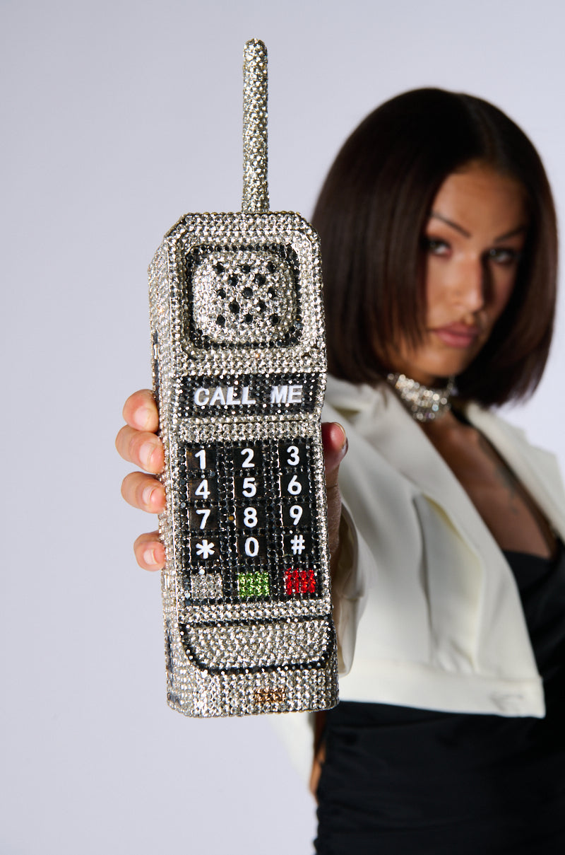 A woman with short dark hair, wearing a white jacket over a black outfit, holds up the CALL ME BLING CLUTCH, a dazzling phone-shaped purse adorned with sparkling rhinestones and featuring the text "CALL ME" on its screen. The background is plain white, keeping the focus on the sparkling accessory. 14