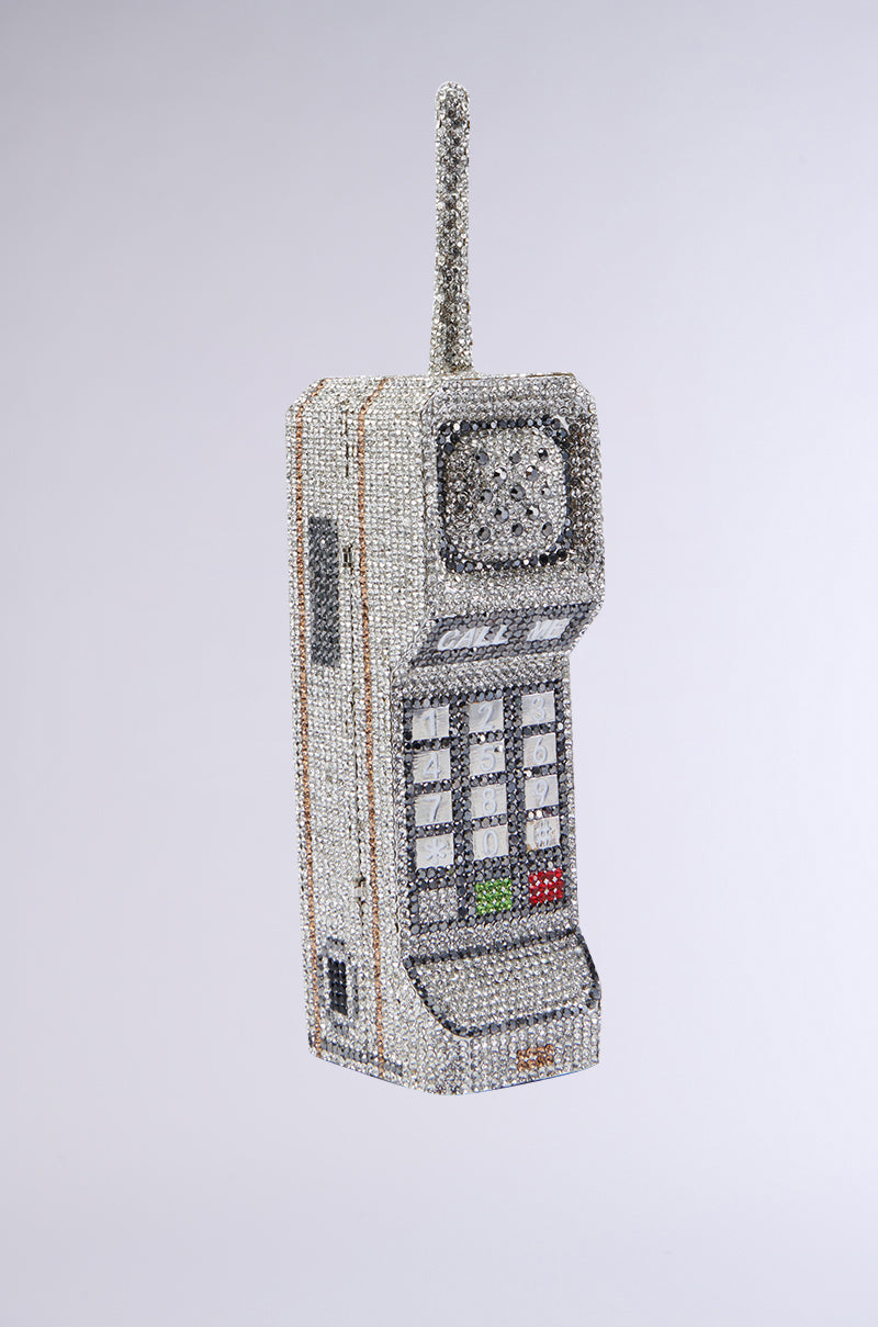 The CALL ME BLING CLUTCH, resembling vintage mobile phone models from the 1980s, is adorned entirely in sparkly rhinestones and features an antenna, numerical keypad, and small screen. It is showcased against a plain grey background. 10