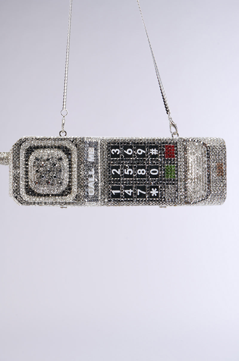 The CALL ME BLING CLUTCH is a sparkling, vintage-inspired phone-shaped purse decorated with rhinestones. It boasts a black numeric keypad with white numbers and symbols, and is draped from a silver chain. The design also includes a rotary dial and antenna, seamlessly merging retro tech aesthetics with contemporary fashion.