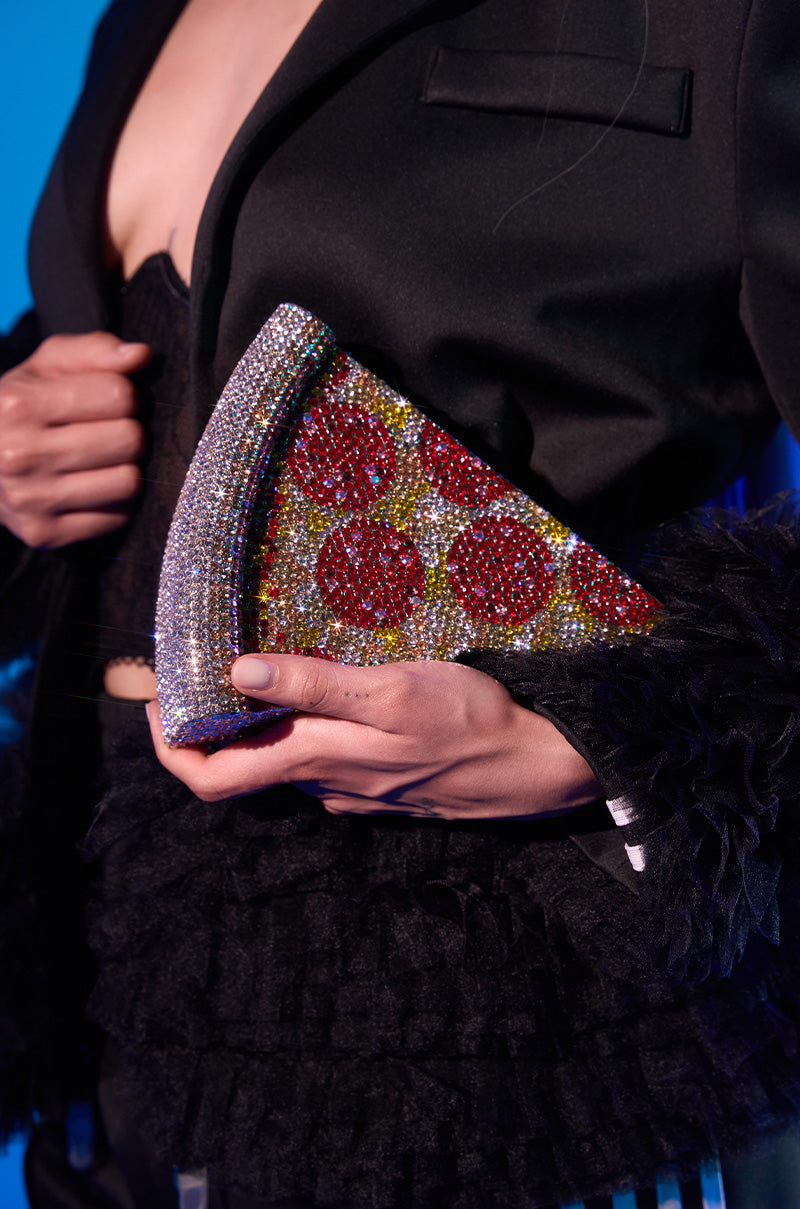 A person in a black ruffled outfit holds the GRAB A SLICE RHINESTONE PIZZA CLUTCH, a sparkling rhinestone handbag shaped like a pepperoni pizza slice, against a blue background.