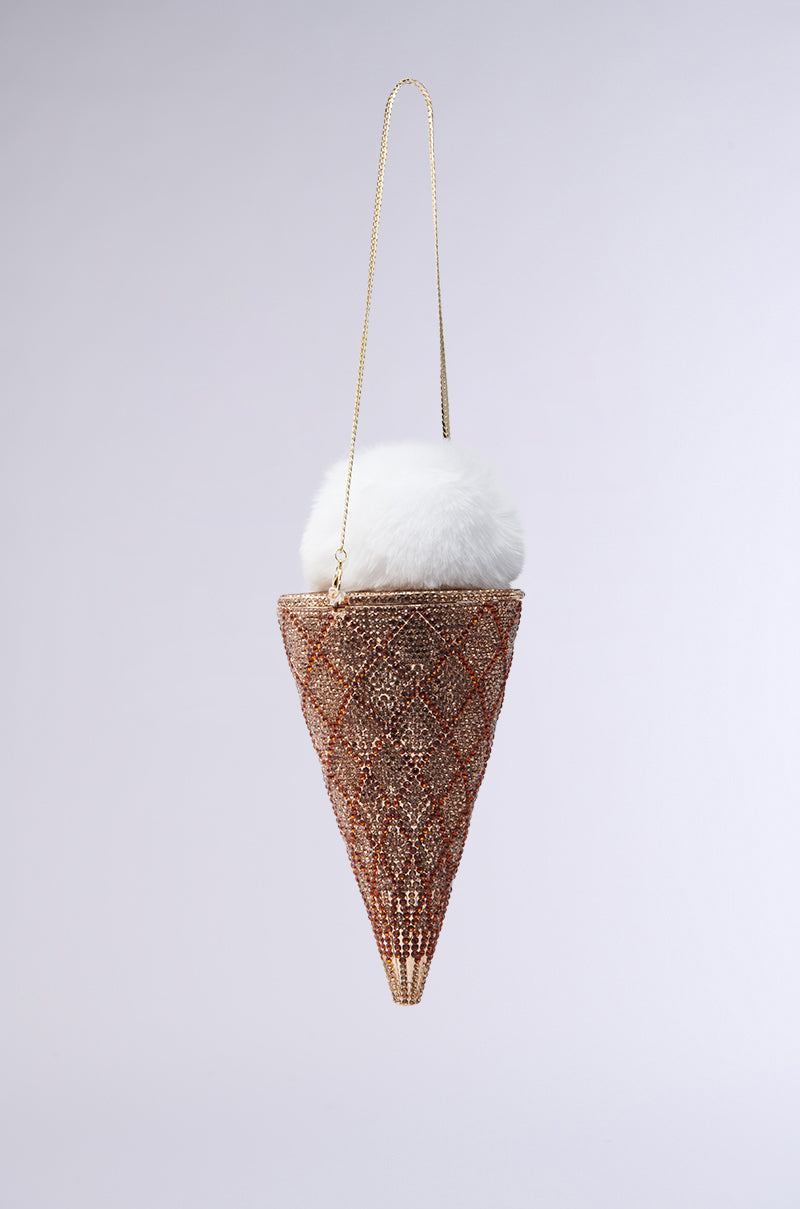 The "LICK IT UP FAUX FUR ICE CREAM BAG" is a decorative accessory shaped like an ice cream cone, featuring a shimmering rhinestone waffle cone topped with a fluffy white pom-pom. A gold chain allows it to hang elegantly against a plain light gray background, enhancing its unique design.