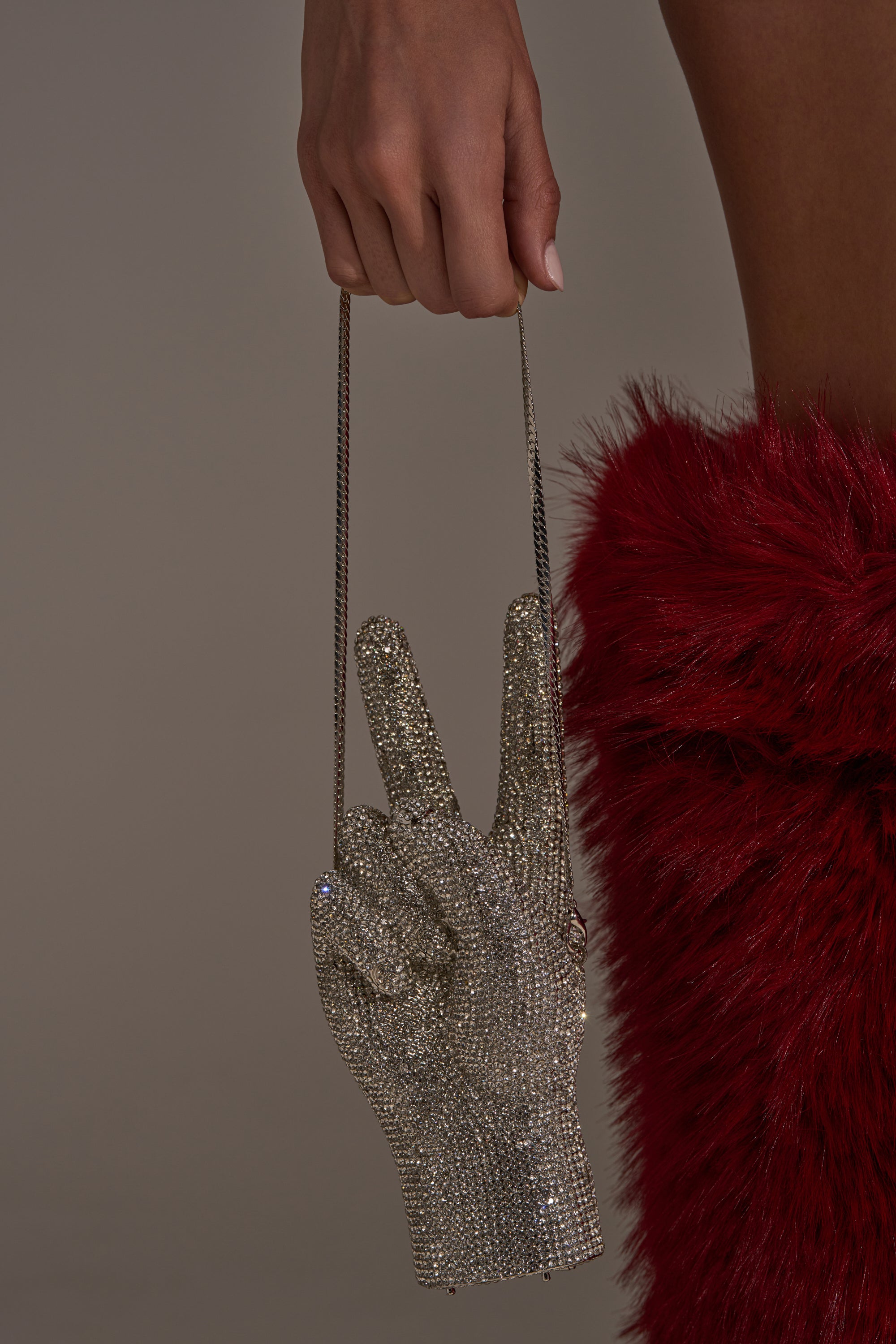 A person holds the PEACE BLING PURSE, a sparkling rhinestone handbag shaped like a peace sign with a thin chain strap, while wearing a red faux fur garment.