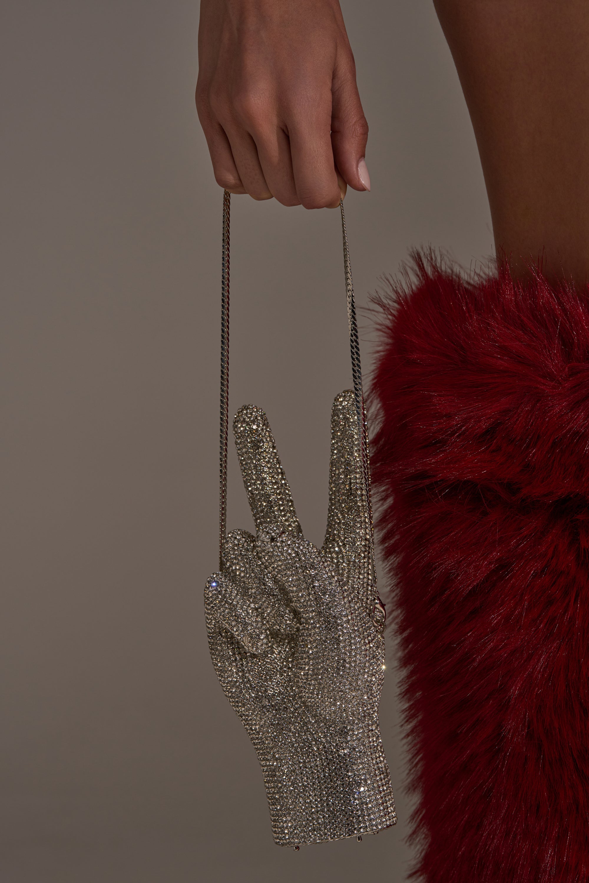 A person holds the PEACE BLING PURSE, a sparkling rhinestone handbag shaped like a peace sign with a thin chain strap, while wearing a red faux fur garment. 5