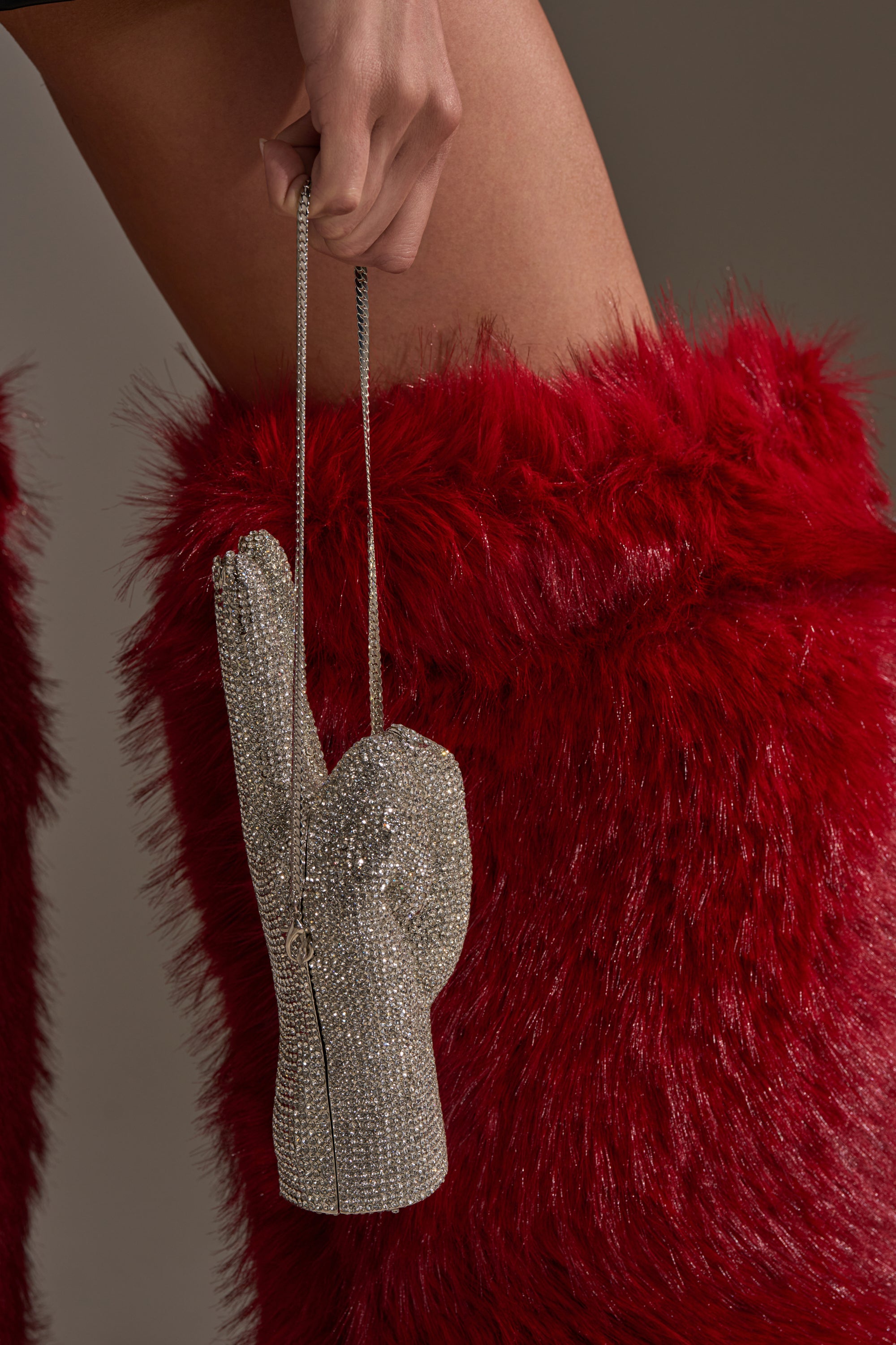 A close-up of a person in tall, bright red furry boots holding the PEACE BLING PURSE—a novelty rhinestone handbag shaped like a hand with a thin chain, adding bold flair to their look.
