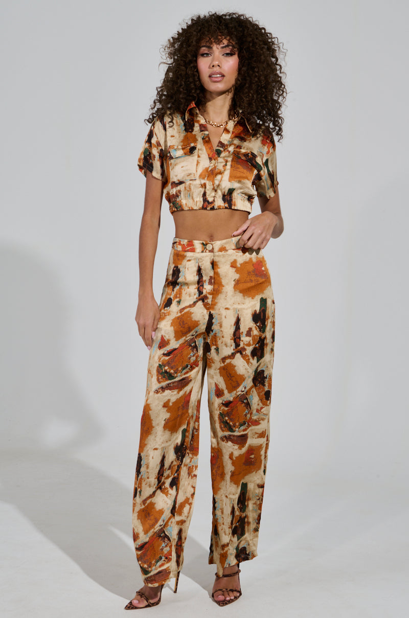 A person with curly hair models a matching outfit featuring a cropped shirt and the CRAZY PANT TROUSER, designed in an abstract brown, orange, and beige pattern. The satin fabrication adds a touch of luxury as they wear high heels and stand against a plain background.