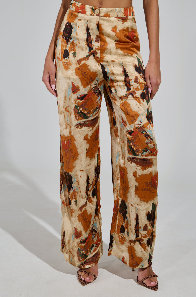 A person models the CRAZY PANT TROUSER with a high-waisted design, showcasing an abstract pattern in shades of orange, brown, and beige. These trousers are paired with open-toed sandals against a simple, light-colored backdrop.