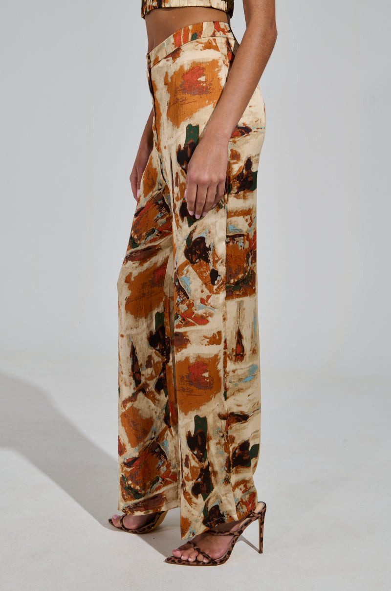Dressed in the CRAZY PANT TROUSER, a person stands in profile, showcasing high-waisted, wide-leg trousers adorned with a colorful abstract pattern featuring shades of brown, orange, and black. They complement this high-rise design with strappy high-heeled sandals.