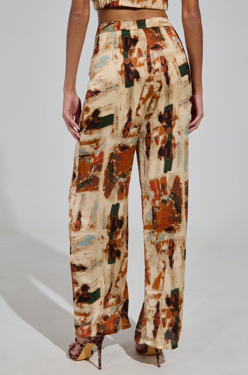 Wearing the CRAZY PANT TROUSER, a person stands modeling its wide-legged, satin design featuring an abstract, multicolored pattern in earthy tones like brown, cream, and teal. The look is completed with high heels against a simple neutral background.