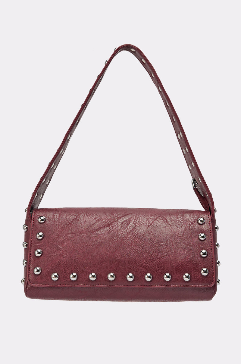 The LOLA STUDDED PURSE is a rectangular maroon handbag with a single strap, featuring silver studs along the edges and strap, displayed on a plain light background. 1