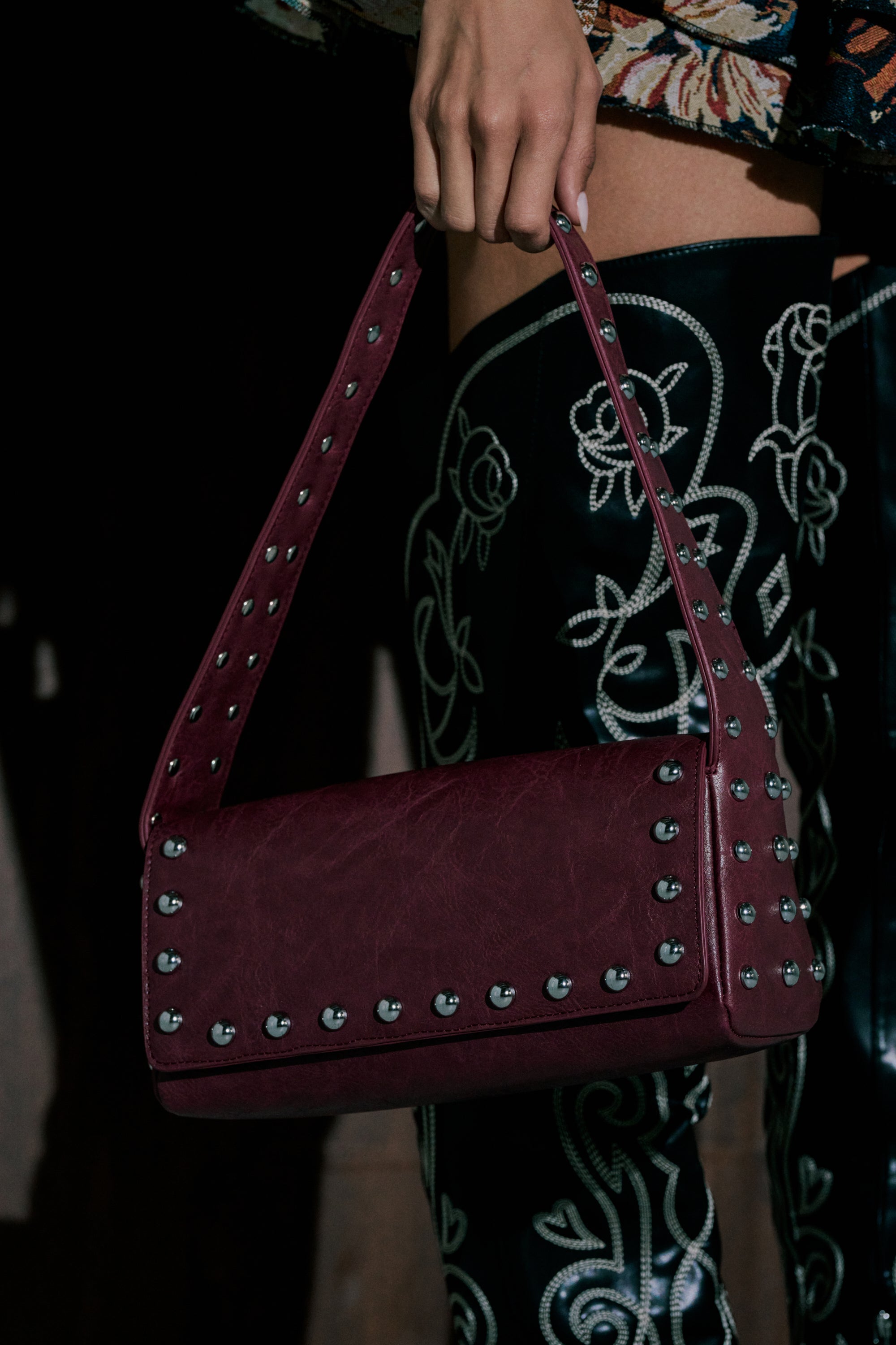 A hand holds the LOLA STUDDED PURSE, a burgundy faux leather handbag with silver studs. Paired with black pants featuring white floral embroidery, it creates a chic Y2K-inspired look. 0
