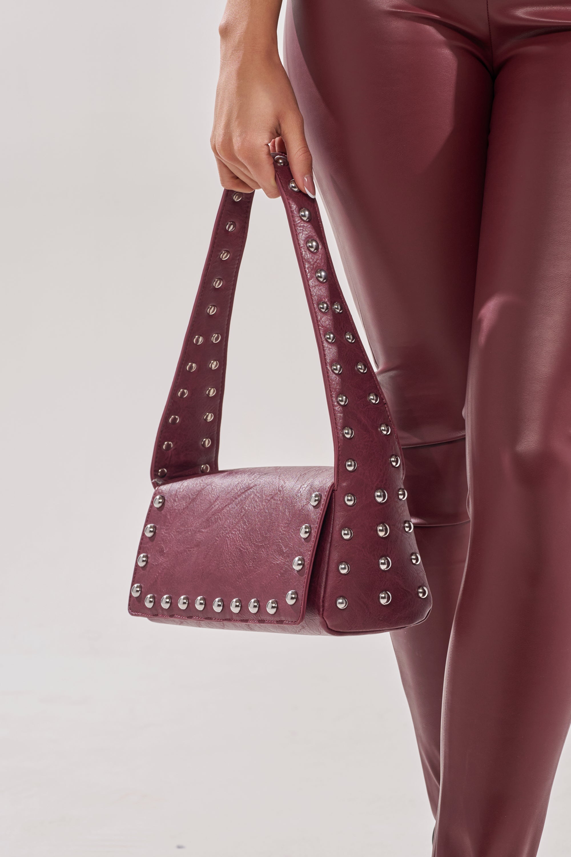 A person in shiny burgundy pants holds the LOLA STUDDED PURSE, a matching burgundy handbag with silver studs on the edges and wide handle, against a plain light background. 5