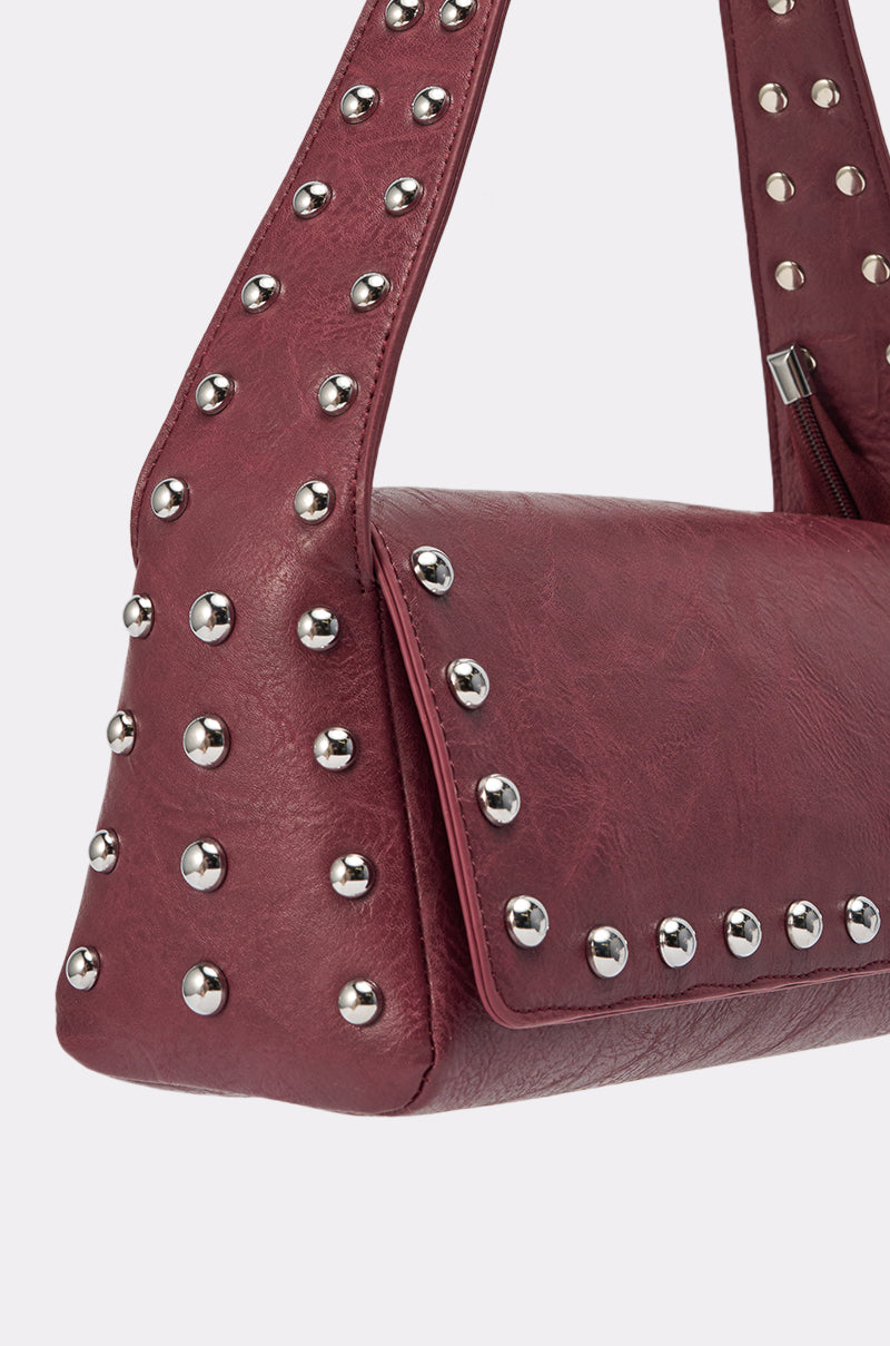 A close-up of the LOLA STUDDED PURSE, a maroon leather handbag featuring large, shiny silver studs on the strap and front flap, displayed against a plain light background. 6