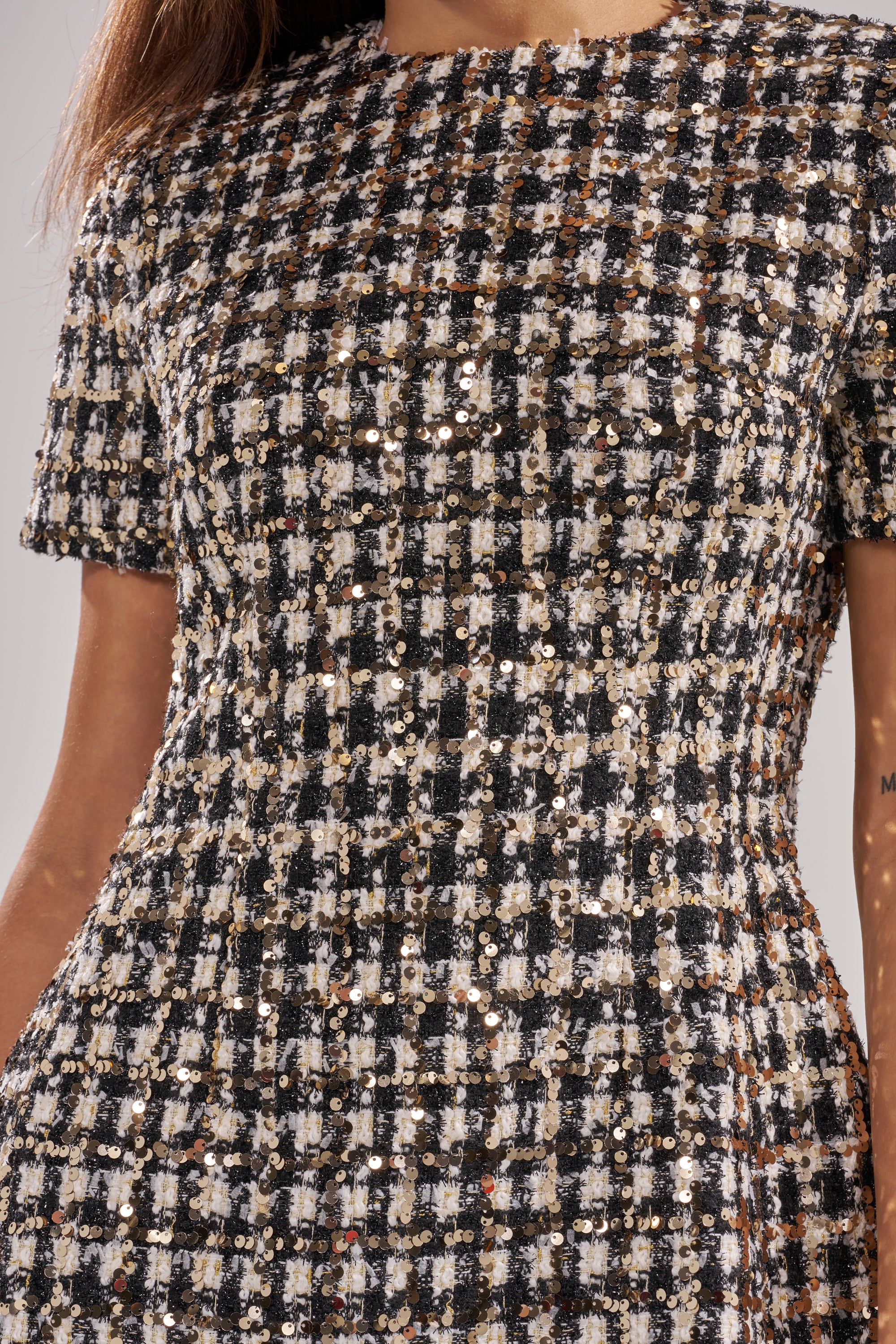 A close-up of a person wearing the GIRLY POP TWEED MINI DRESS, featuring a black, white, and gold sequin plaid pattern with short sleeves; their head is out of frame and arms are at their sides.