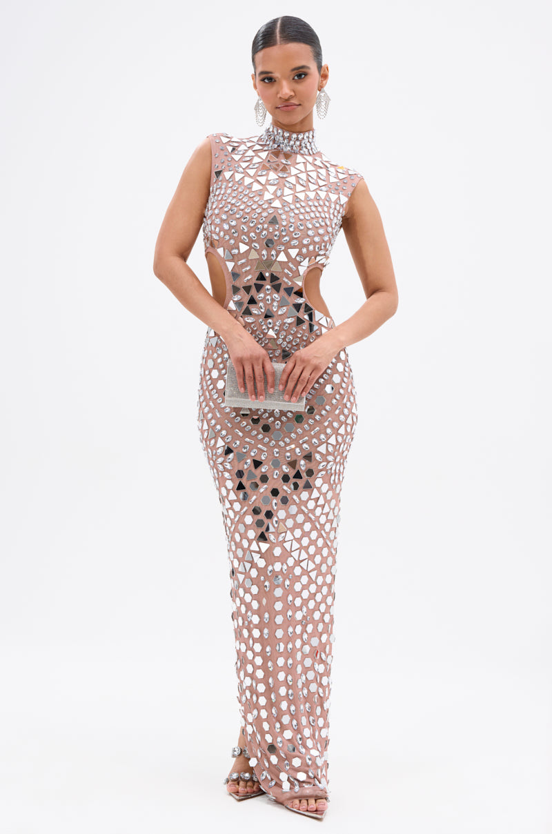 Woman wearing the QUEEN OF EVERYTHING MIRRORED MAXI DRESS IN SILVER, a sleeveless, floor-length gown with geometric cutouts and metallic accents, holding a silver clutch against a plain white background.