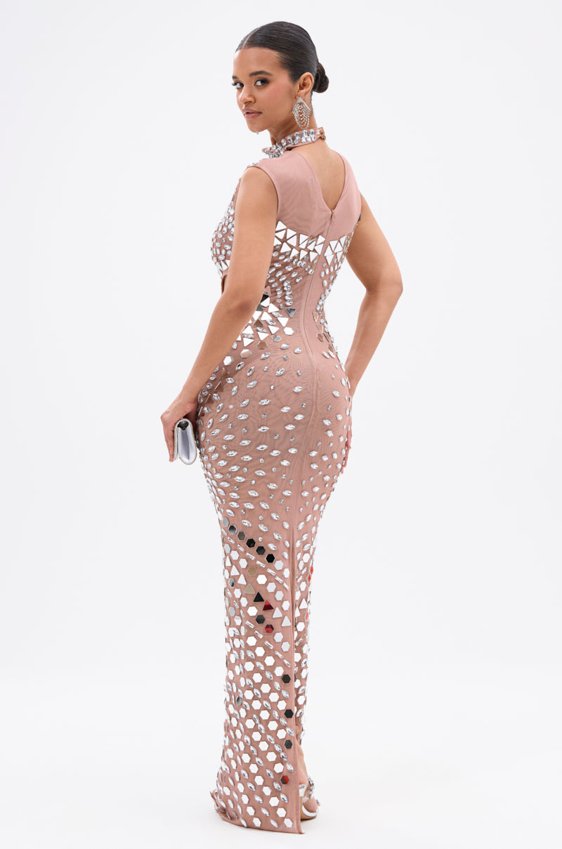 A woman wears the QUEEN OF EVERYTHING MIRRORED MAXI DRESS IN SILVER, featuring silver circular cutouts. She holds a clutch, looks over her shoulder, and accessorizes with a sleek bun, large earrings, and a statement necklace on a white background.