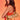 A woman with long, wavy brown hair poses against an orange background, wearing a red and orange dress and holding the TOO ROYAL CROSS PURSE—a metallic gold purse—over her shoulder.