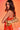 A woman with long, wavy brown hair poses against an orange background, wearing a red and orange dress and holding the TOO ROYAL CROSS PURSE—a metallic gold purse—over her shoulder.