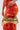 A person in a fitted red-orange patterned dress holds the TOO ROYAL CROSS PURSE—a metallic gold bag with textured details and a jeweled cross-shaped clasp—against a plain background.