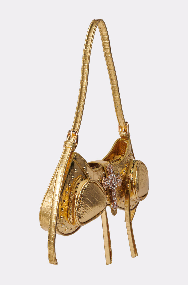 The TOO ROYAL CROSS PURSE is a gold handbag with a crocodile texture, two rounded compartments, jeweled cross detail on the front, single shoulder strap, and studded accents. 3