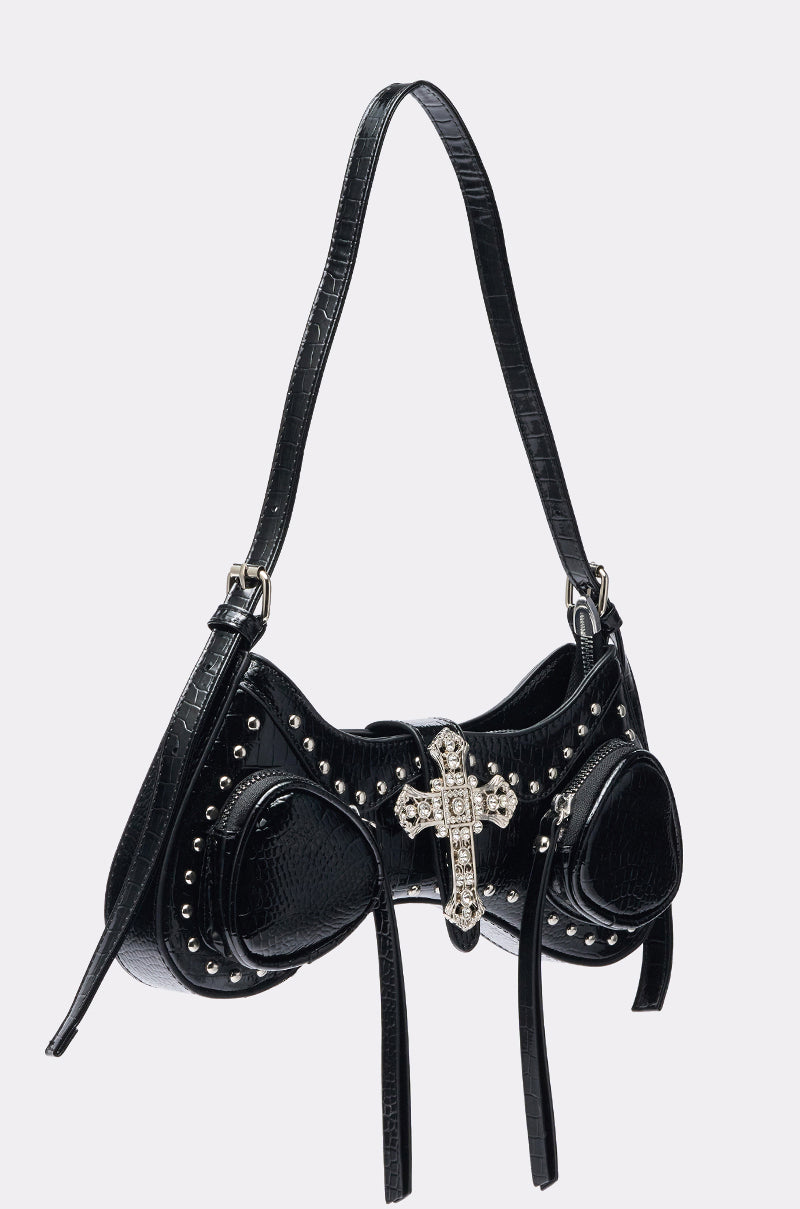 The HEAVEN SENT PURSE is a chic black patent faux leather bag designed like a bra, with silver studs, two pouch-like cups, long straps, and a large jeweled cross centerpiece. The handle attaches to both sides for stylish carrying. 2