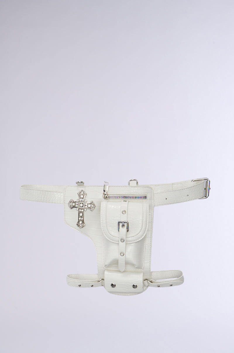 The CROSSING THE LINE PU BELT BAG IN WHITE is a faux leather accessory with an ornate silver cross embellishment. It features a buckle and zipper closure front pocket. Its minimalist and modern design pairs well with a plain light background.