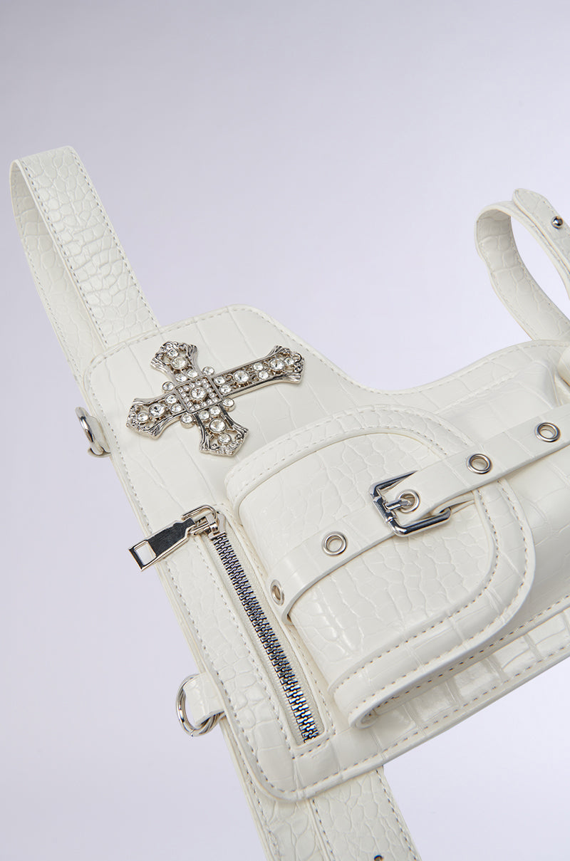 The CROSSING THE LINE PU BELT BAG IN WHITE is a faux leather bag with a chic crocodile pattern and silver hardware. It boasts a rhinestone cross, zipper detail, buckle strap, and multiple loops. The neutral backdrop enhances this elegant blend of fashion and function.