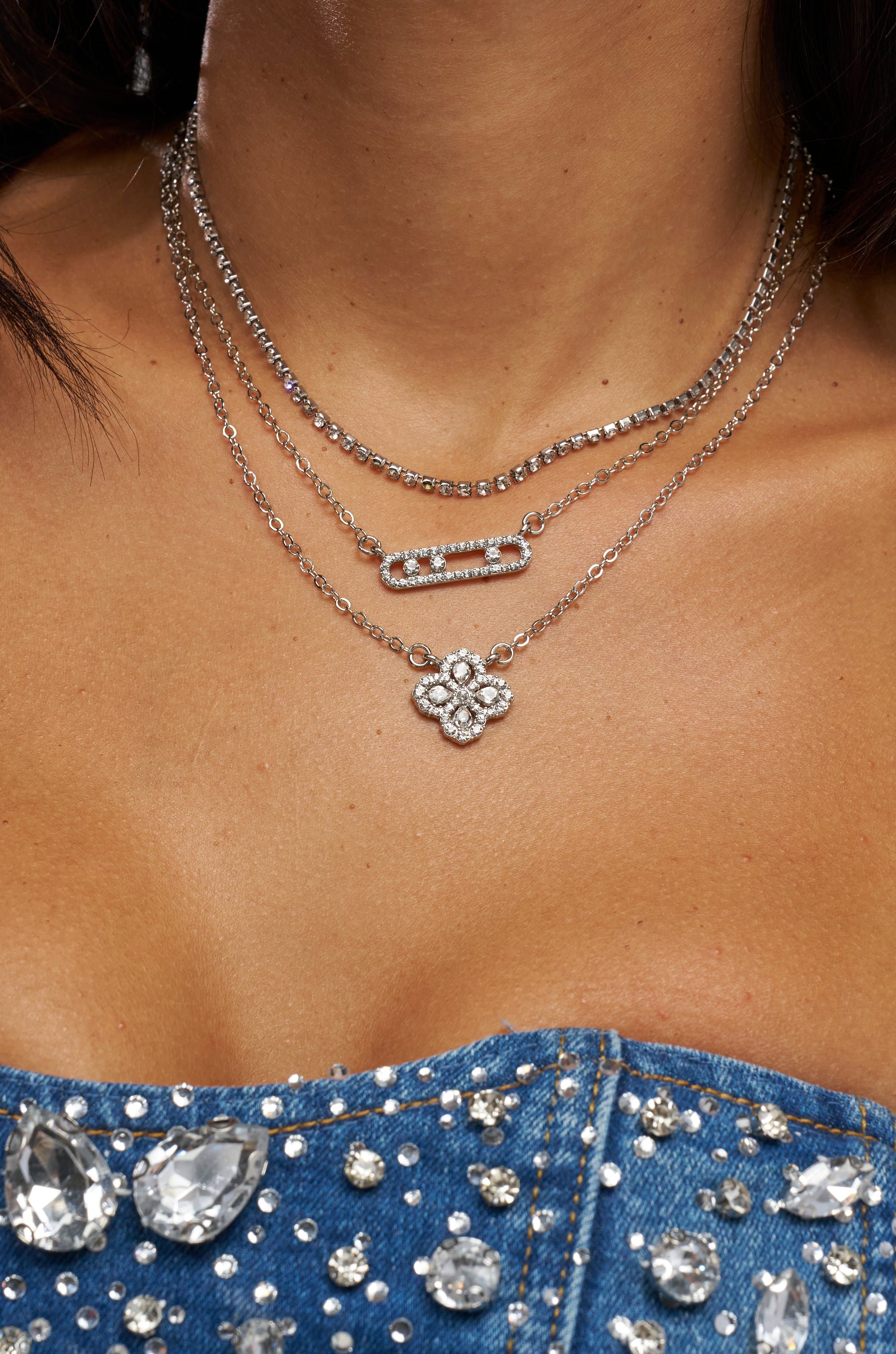 A close-up of a woman wearing the LUXE LIFE NECKLACE, a layered silver set with floral and rectangular bar pendants, styled with a denim top decorated with large rhinestones.
