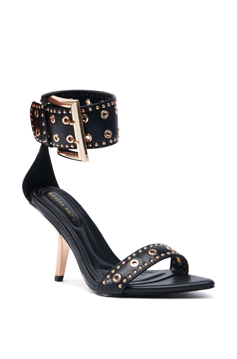 The AZALEA WANG LOBELIA sandal is a black faux leather stiletto with sculpted heels and decorative studs on the ankle and toe straps. It features a gold buckle, black hardware embellishments, and a sleek sole.