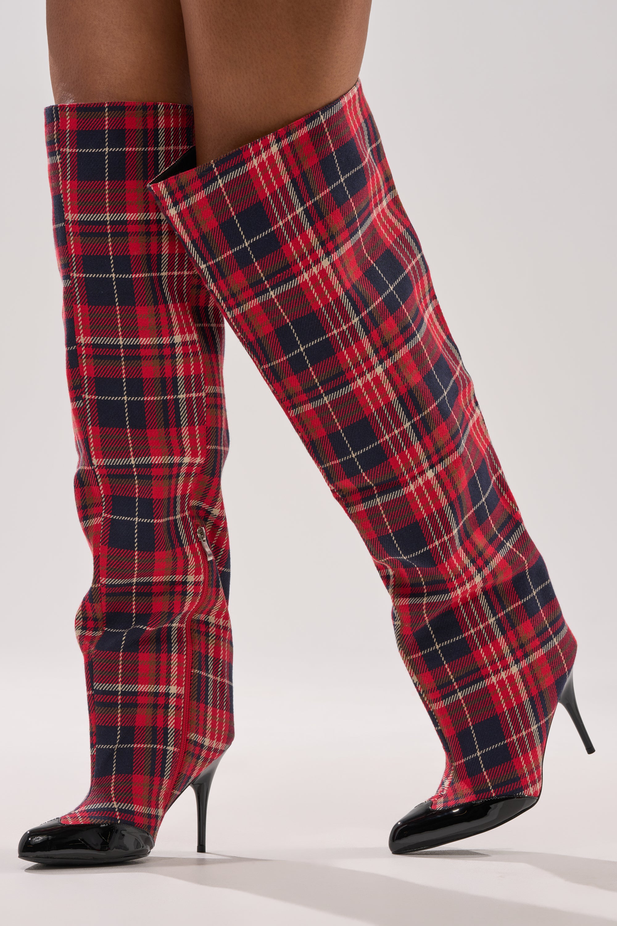 A person stands against a plain background with one leg slightly in front of the other, wearing the AKIRA GOLD LOCHANA MULTI PLAID FLANNEL SEXY BOOT—tall, pointed-toe, over-the-knee boots with stiletto heels.