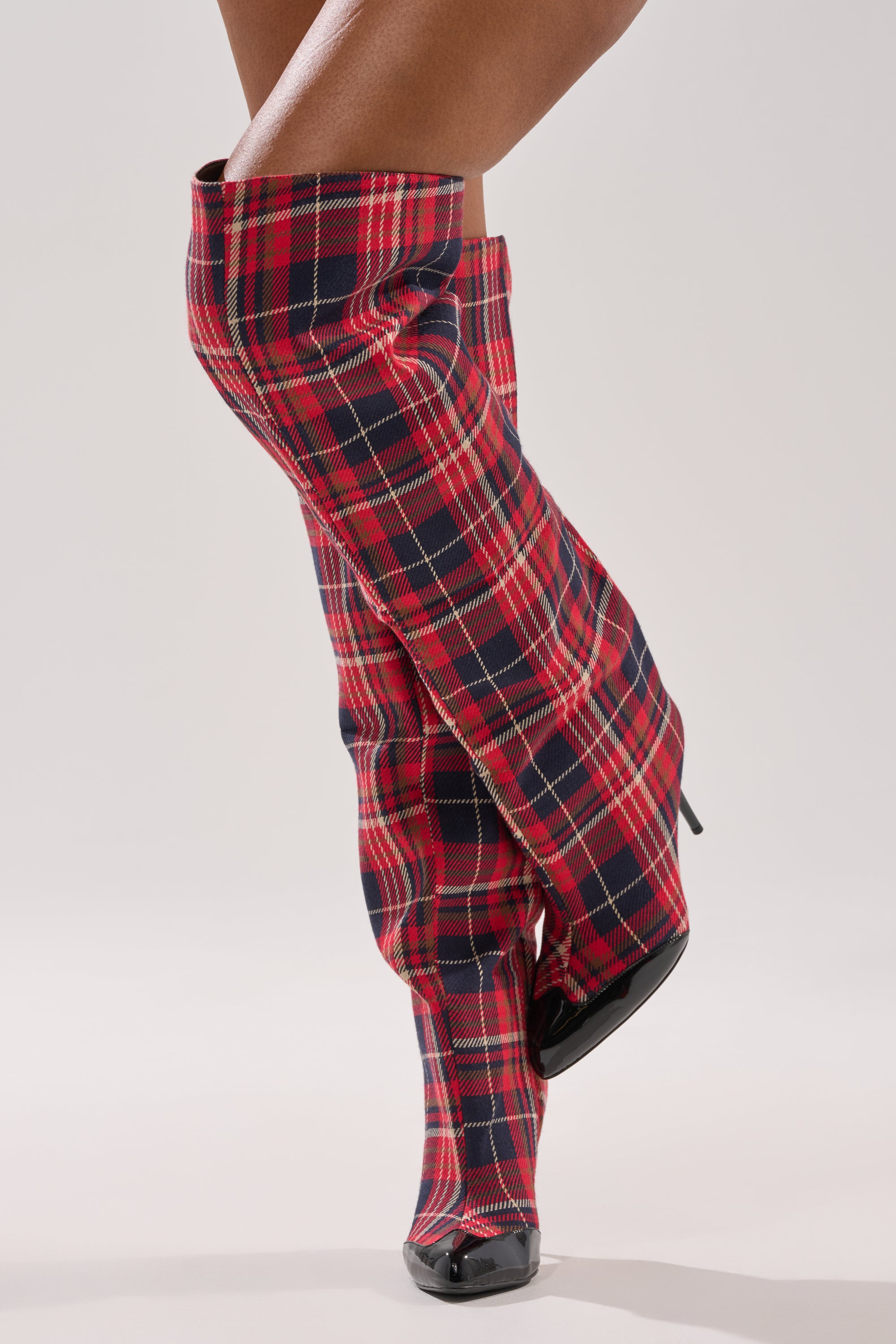 A person crosses their legs against a plain light gray background, wearing AKIRA GOLD LOCHANA Multi Plaid Flannel Sexy Boots with stiletto heels.
