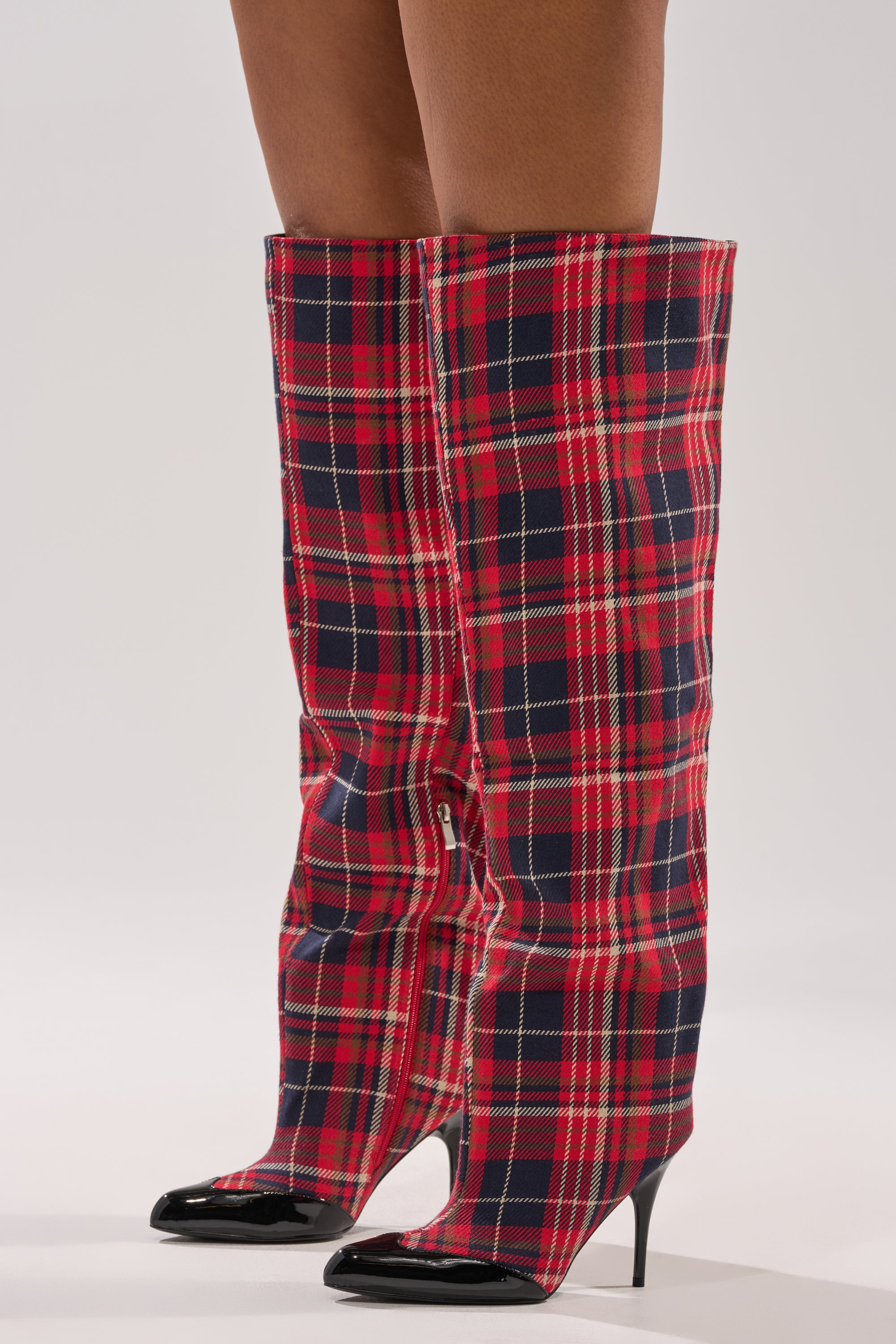 A person models the AKIRA GOLD LOCHANA MULTI PLAID FLANNEL SEXY BOOT, featuring over-the-knee plaid flannel design and black pointed stiletto heels, standing on a white background.