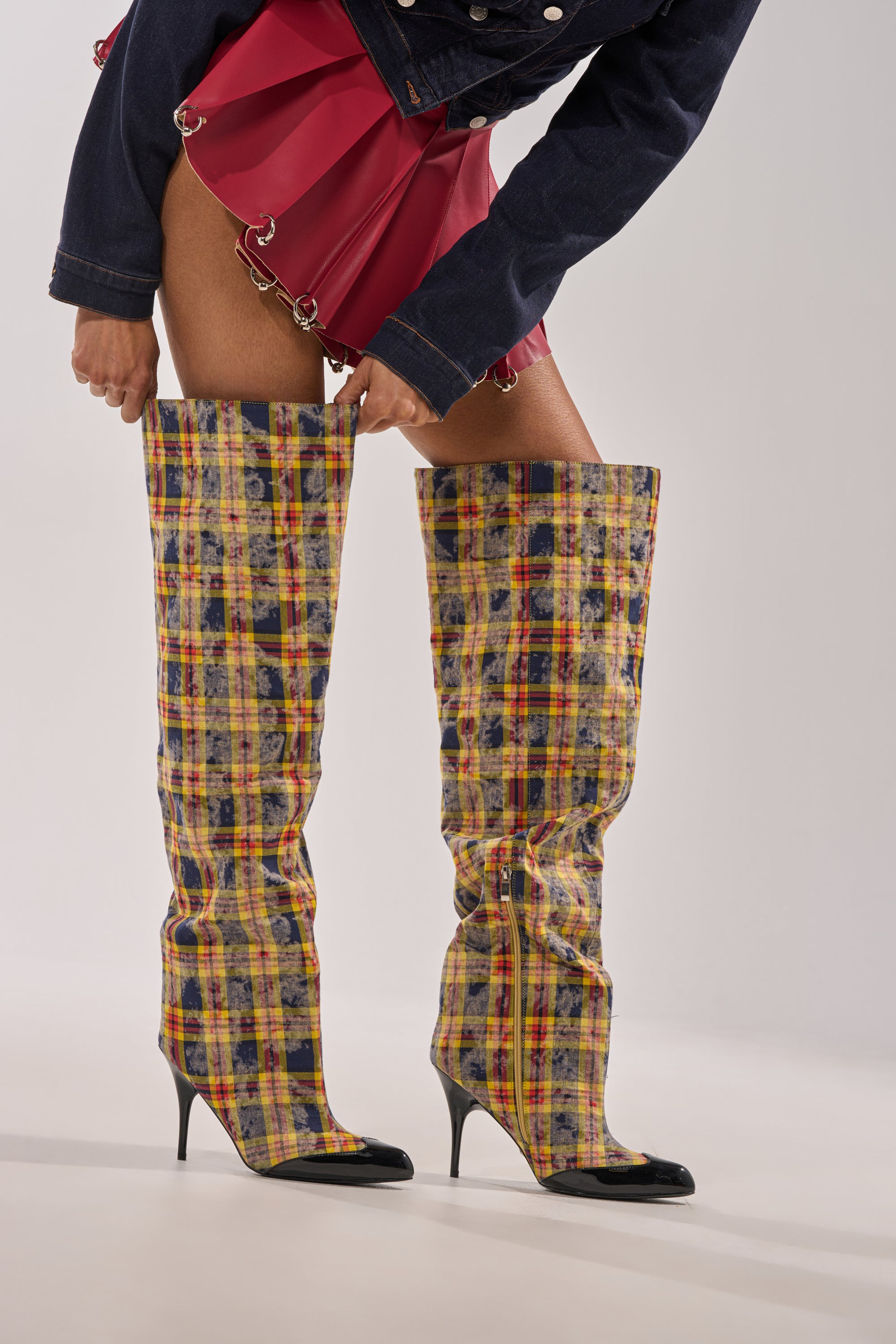 Wearing a short red pleated skirt, dark jacket, and AKIRA WANG LOCHANA MULTI YELLOW PLAID FLANNEL SEXY BOOTS, a person bends forward to adjust the boots. Only the lower half of their body is shown. 1