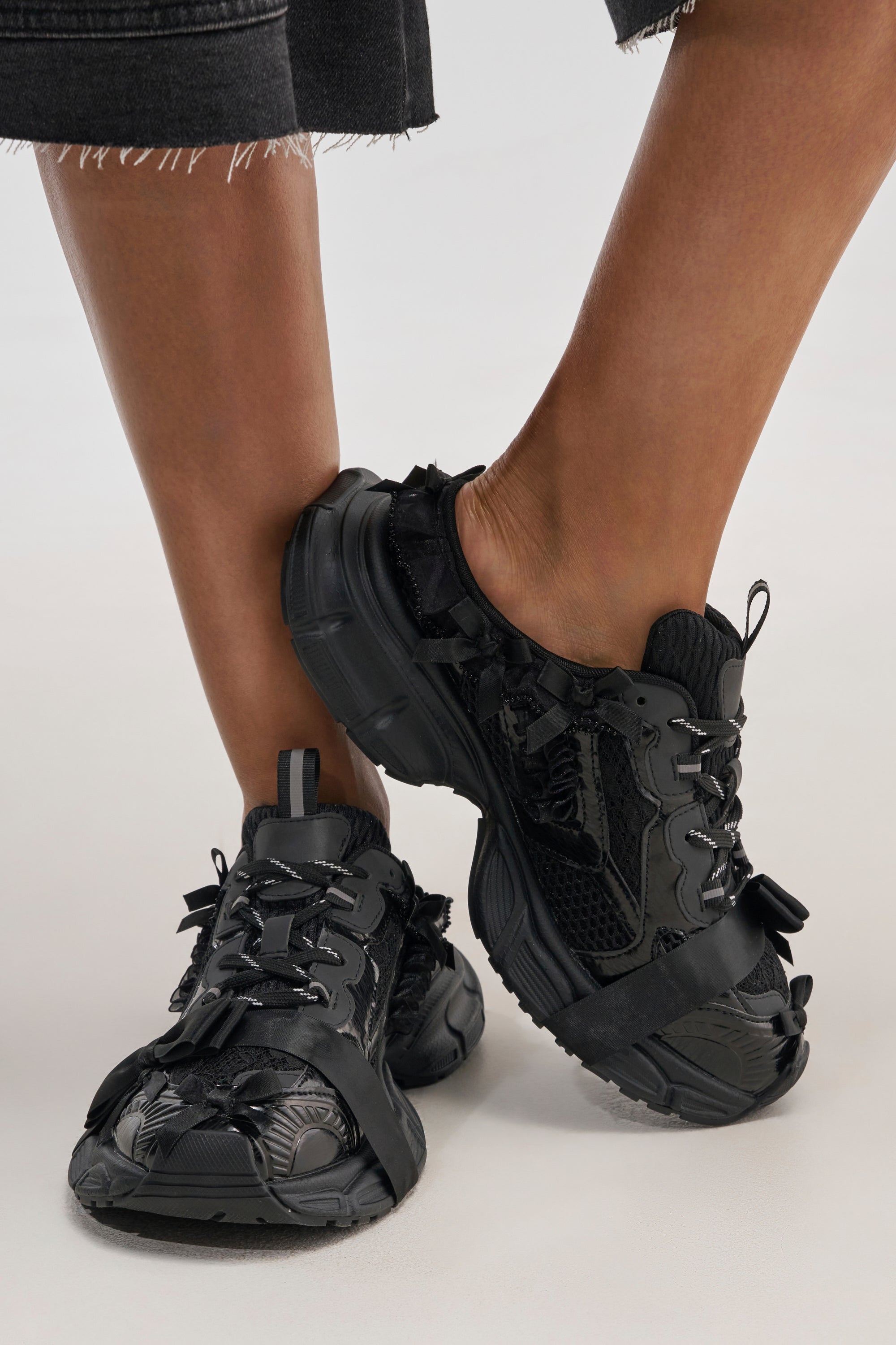 Close-up of a person wearing the AKIRA GOLD LOCKET BLACK SLIP ON SNEAKER, featuring textured details and black-and-white laces. The person stands with legs crossed at the ankles, paired with dark, frayed-hem pants.