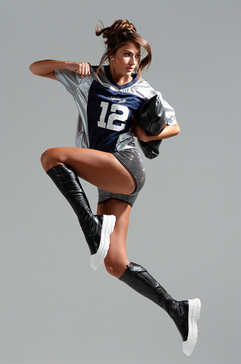 A woman is mid-jump against a gray background, showcasing her streetwear style. She sports knee-high AZALEA WANG LOIS 4 WAY STRETCH MID CALF FLATFORM BOOTS IN BLACK, gray and black checkered shorts, and a sports jersey with the number 12. Her hair is up, and she holds a black jacket draped over her arm.