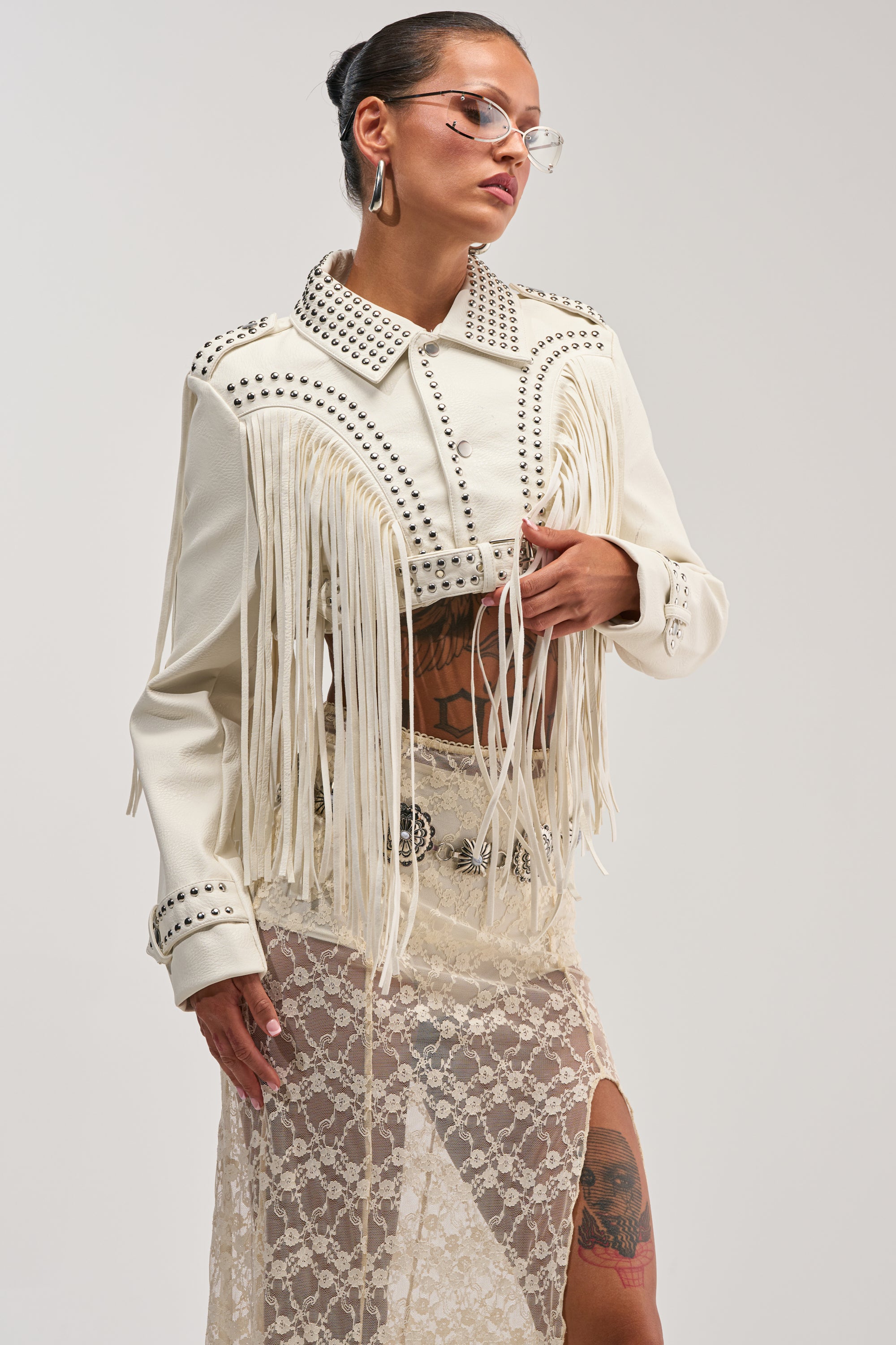 Wearing the RUMI CROPPED MOTO IN WHITE, a person stands confidently against a plain background. The white faux leather fringe jacket with silver studs is paired with a sheer lace skirt, clear sunglasses, and large earrings. Tattoos are visible on their legs and chest.