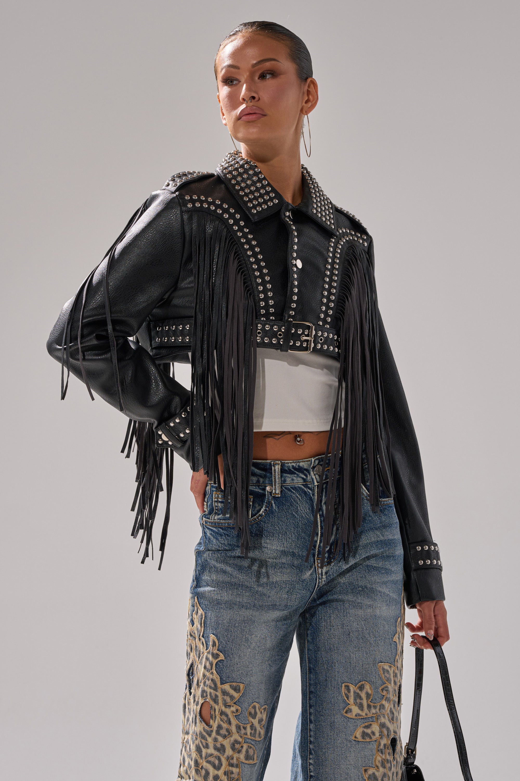 Wearing the RUMI CROPPED MOTO— a black studded cropped moto jacket with fringe—over a white crop top and beige-embroidered blue jeans, a person stands holding a black handbag, confidently gazing to the side against a plain background. 0