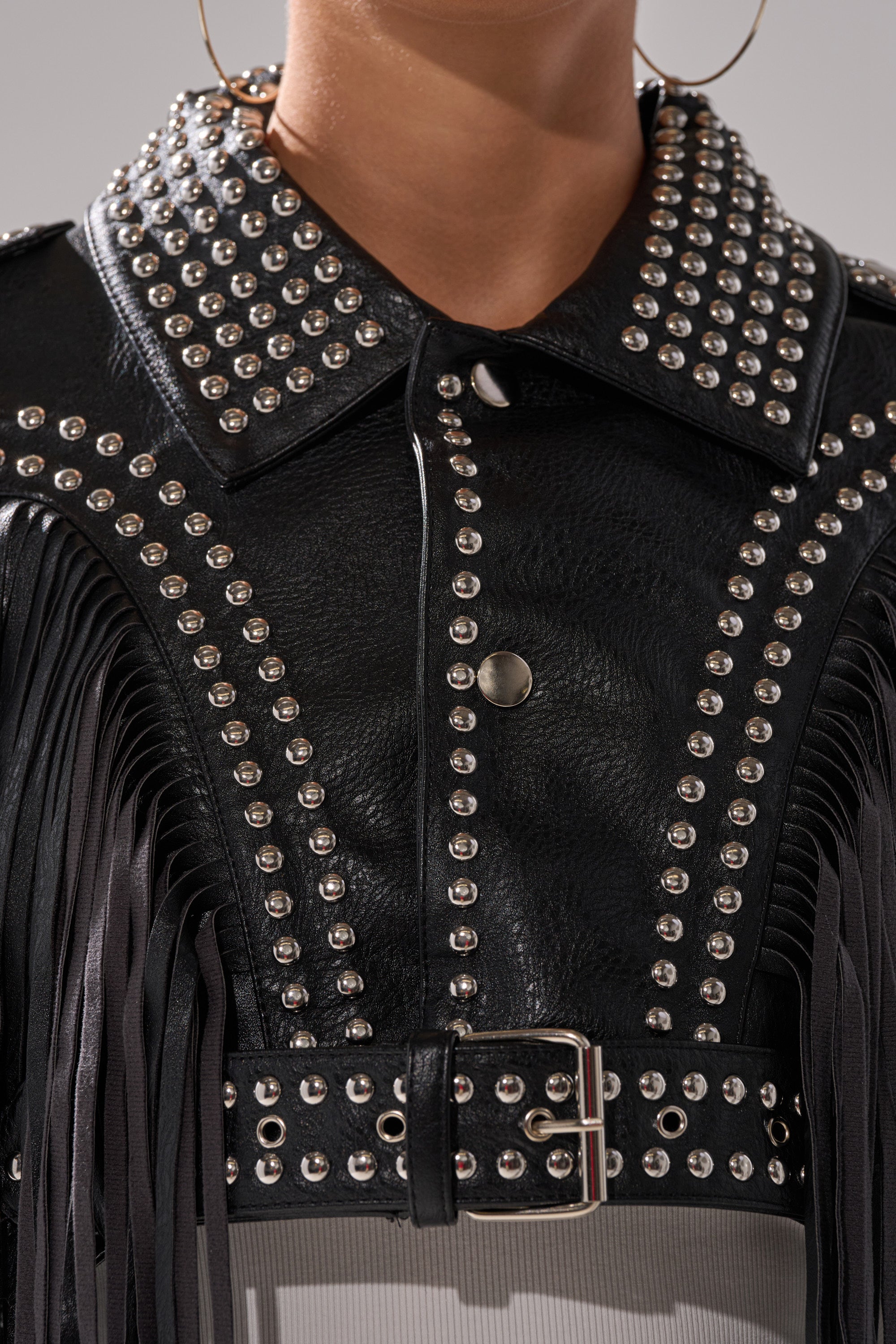 Close-up of the RUMI CROPPED MOTO—a black fringe moto jacket with silver studs, pointed collar, metal buttons, buckle belt, and large hoop earrings worn by the model. 9