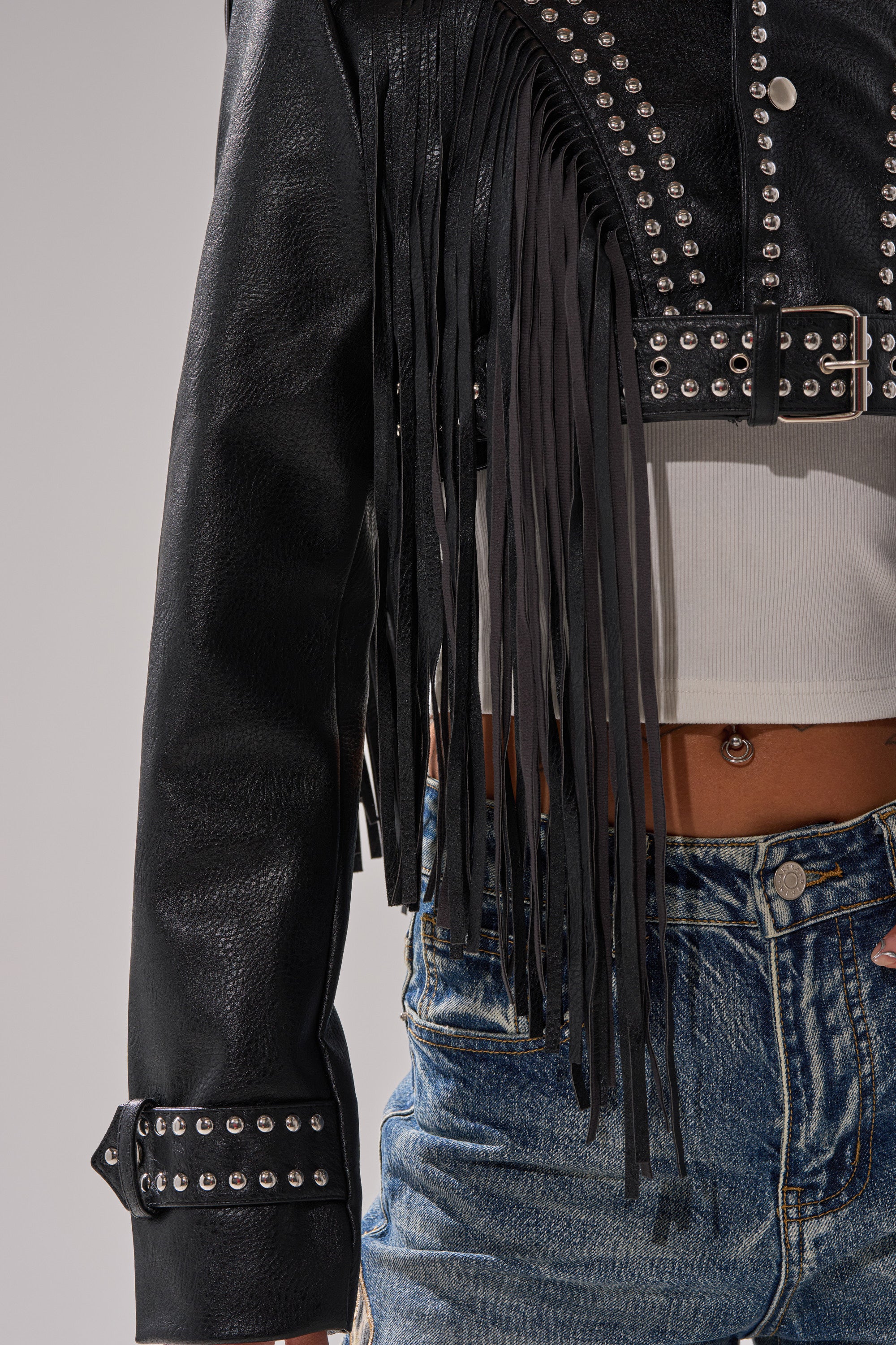 A person wears the RUMI CROPPED MOTO black fringe jacket styled as a cropped moto, paired with a white crop top and blue denim jeans. The image highlights the jacket details and a belly button piercing, focusing on the torso. 10