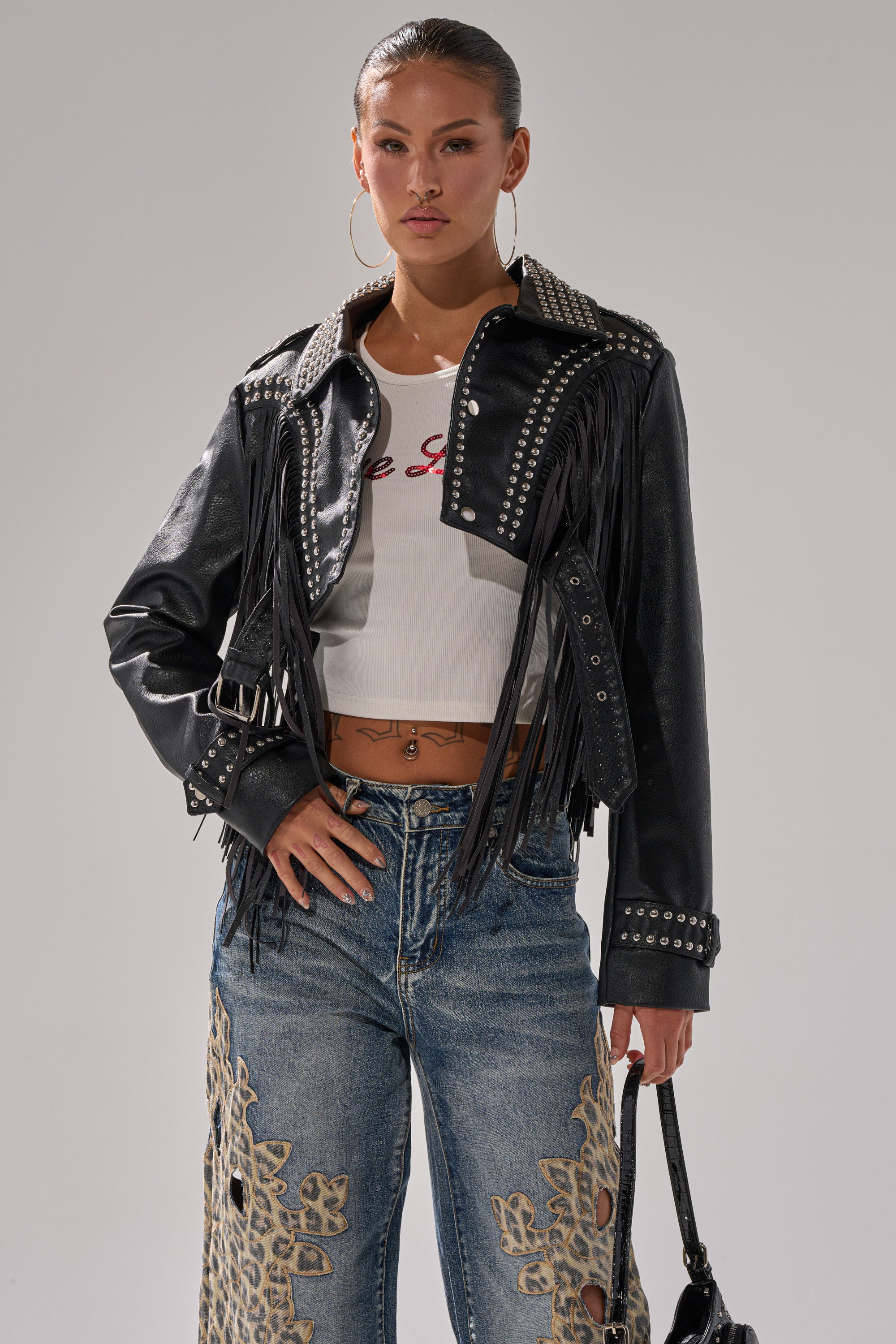 A woman wears the RUMI CROPPED MOTO black faux leather fringe jacket over a white graphic crop top and blue embroidered jeans, holding a black handbag and posing confidently against a plain background. 1
