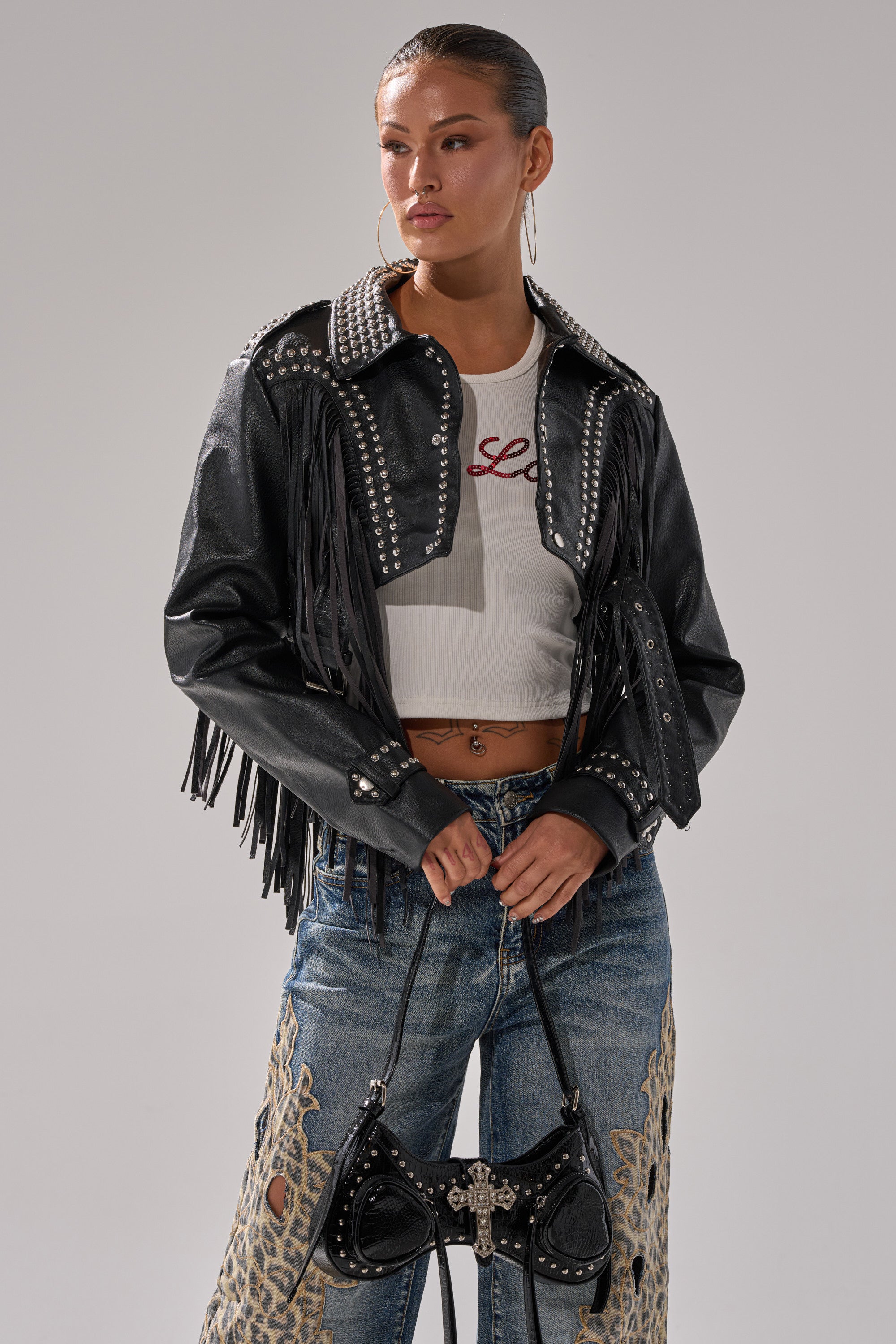 A woman stands confidently in the RUMI CROPPED MOTO black jacket with fringe and studs, a white crop top, distressed jeans with lace accents, and carries a black purse featuring a metallic cross embellishment. 2
