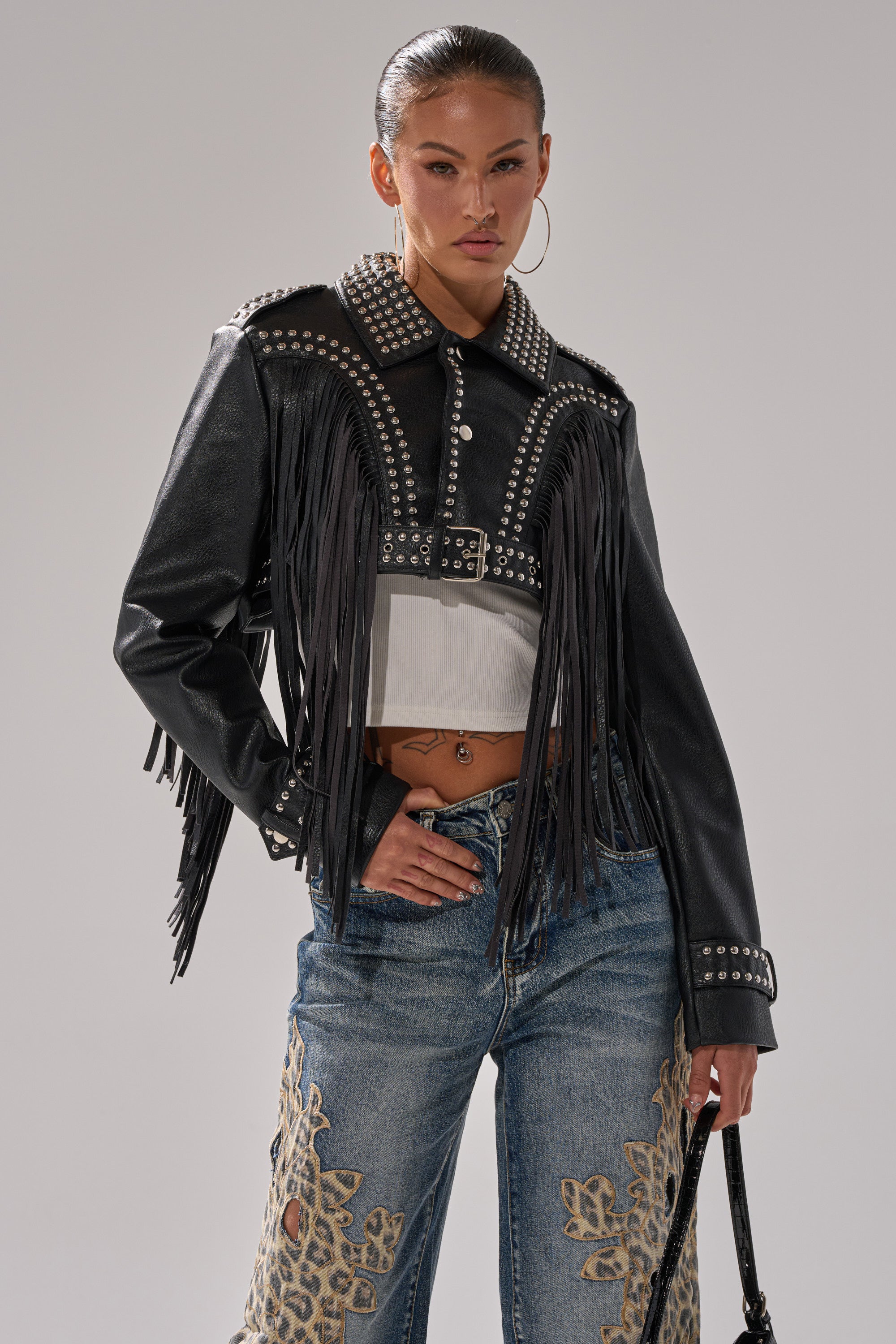 A woman poses in the RUMI CROPPED MOTO, a black fringe moto jacket with studs, styled over a white crop top and blue embroidered jeans, accessorized with a black bag and large hoop earrings. 3