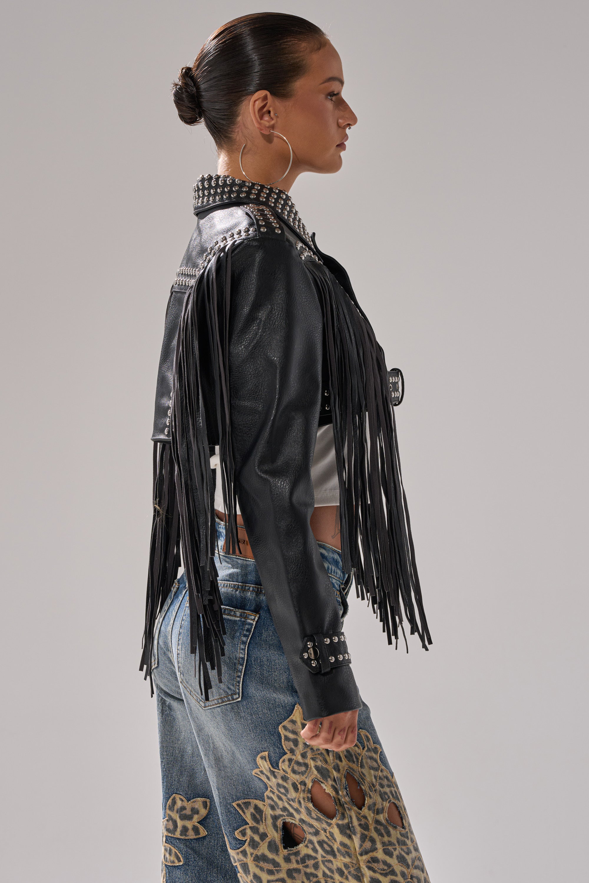 A woman in profile wears the RUMI CROPPED MOTO jacket with fringe and studded details over a white top and patterned cutout blue jeans. Her hair is tied back in a neat bun, and she accessorizes with hoop earrings. 4
