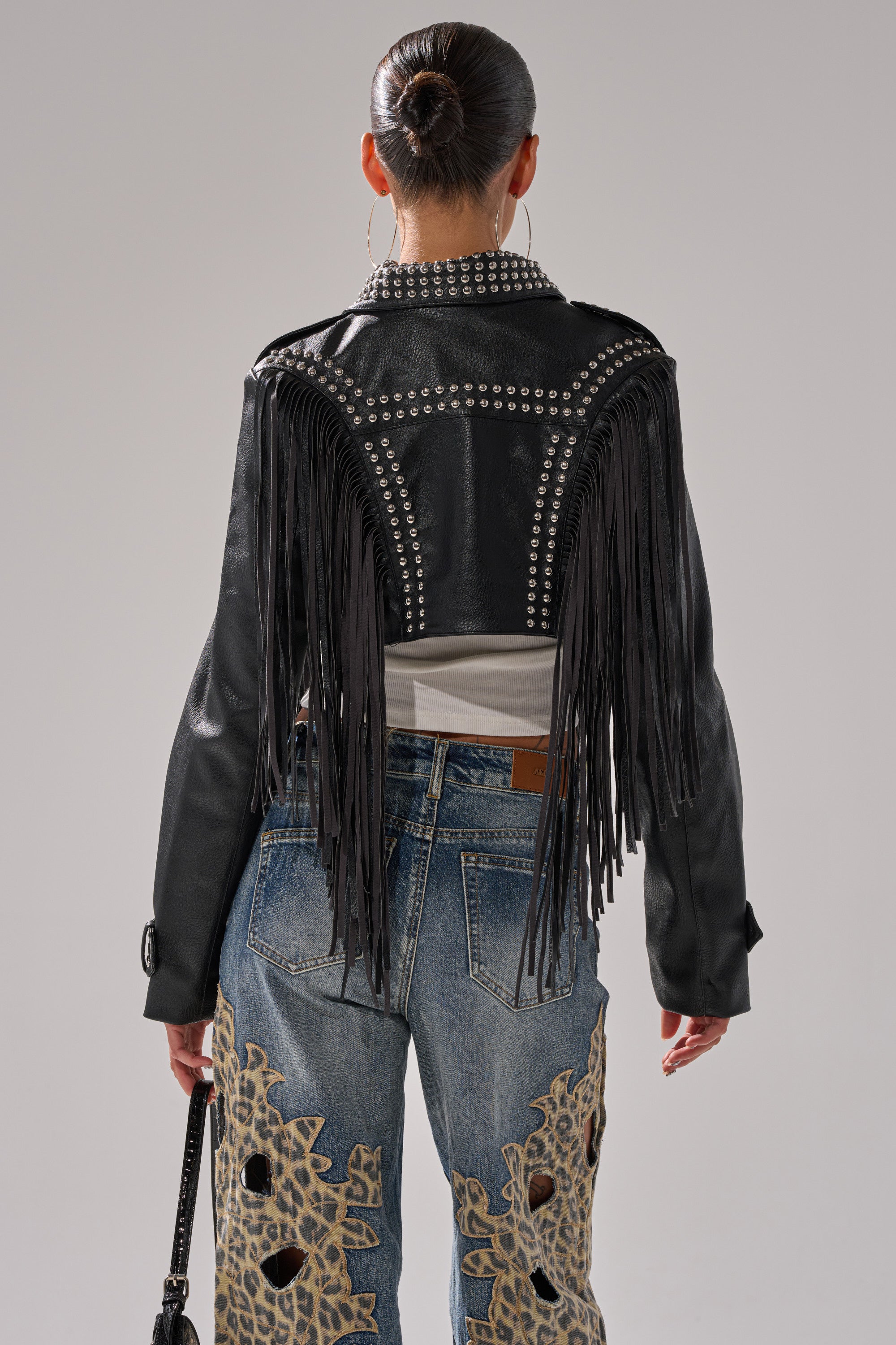 A person with their hair in a bun wears the RUMI CROPPED MOTO, a black fringe jacket with silver studs, paired with blue jeans featuring bold tan lace-like appliqué, adding an edgy touch as they face away from the camera. 5