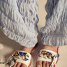 Close-up of a person in light blue ruched wide-leg pants, wearing the AKIRA LORCAN WHITE SANDAL with vegan leather, metallic gold chain, and colorful patterned fabric straps, standing on a light surface.