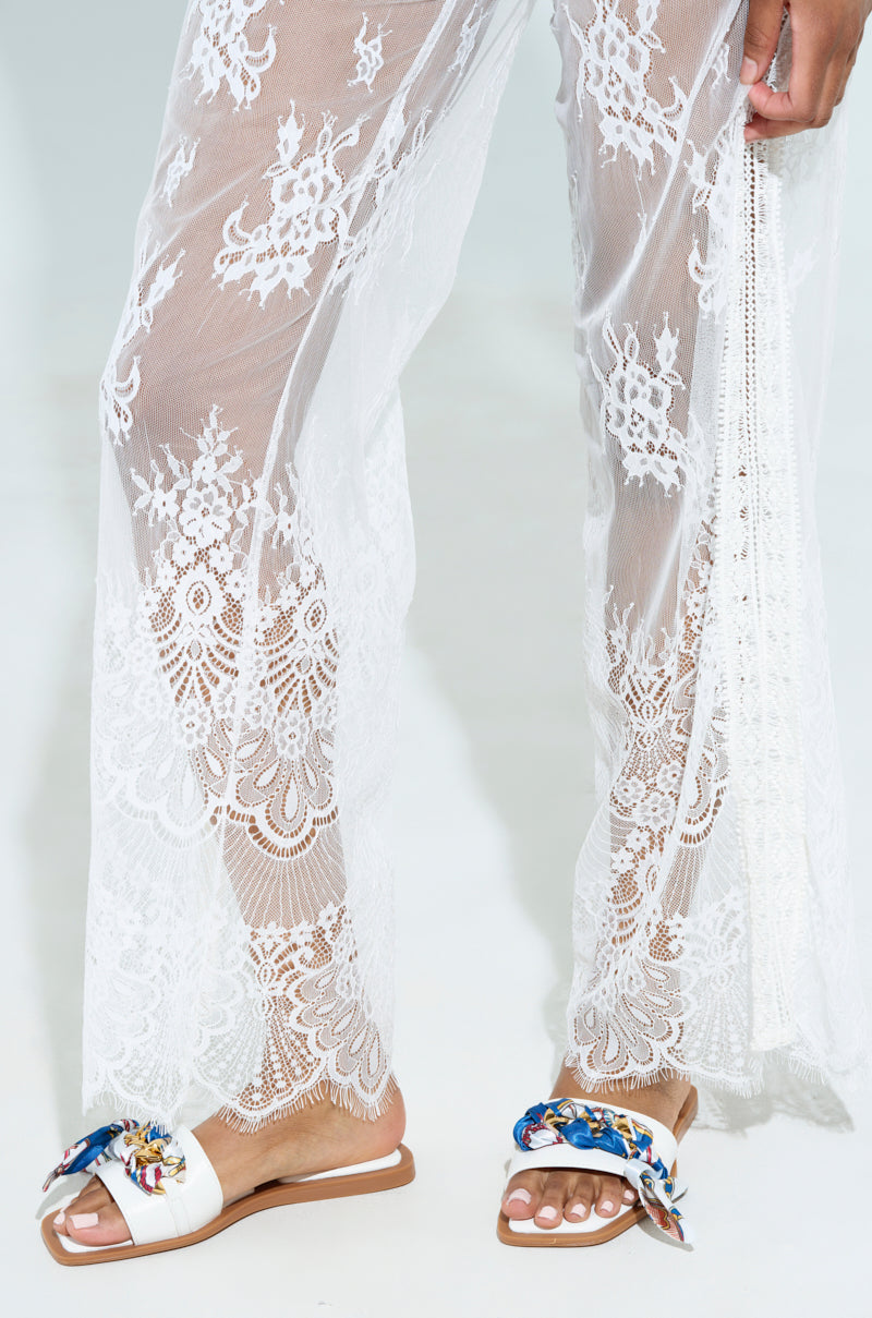 Wearing sheer white lace pants against a light backdrop, this individual dons the AKIRA LORCAN WHITE SANDAL. These slide-on flats feature vegan leather with colorful fabric details. The floral-patterned lace enhances the ensemble beautifully.