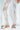 Wearing sheer white lace pants against a light backdrop, this individual dons the AKIRA LORCAN WHITE SANDAL. These slide-on flats feature vegan leather with colorful fabric details. The floral-patterned lace enhances the ensemble beautifully.