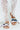 A close-up highlights a person's feet in AKIRA LORCAN WHITE SANDALS, featuring colorful scarf accents and a metallic gold chain. Paired with long white lace pants, these chic slide-on sandals stand out against a light, neutral background.