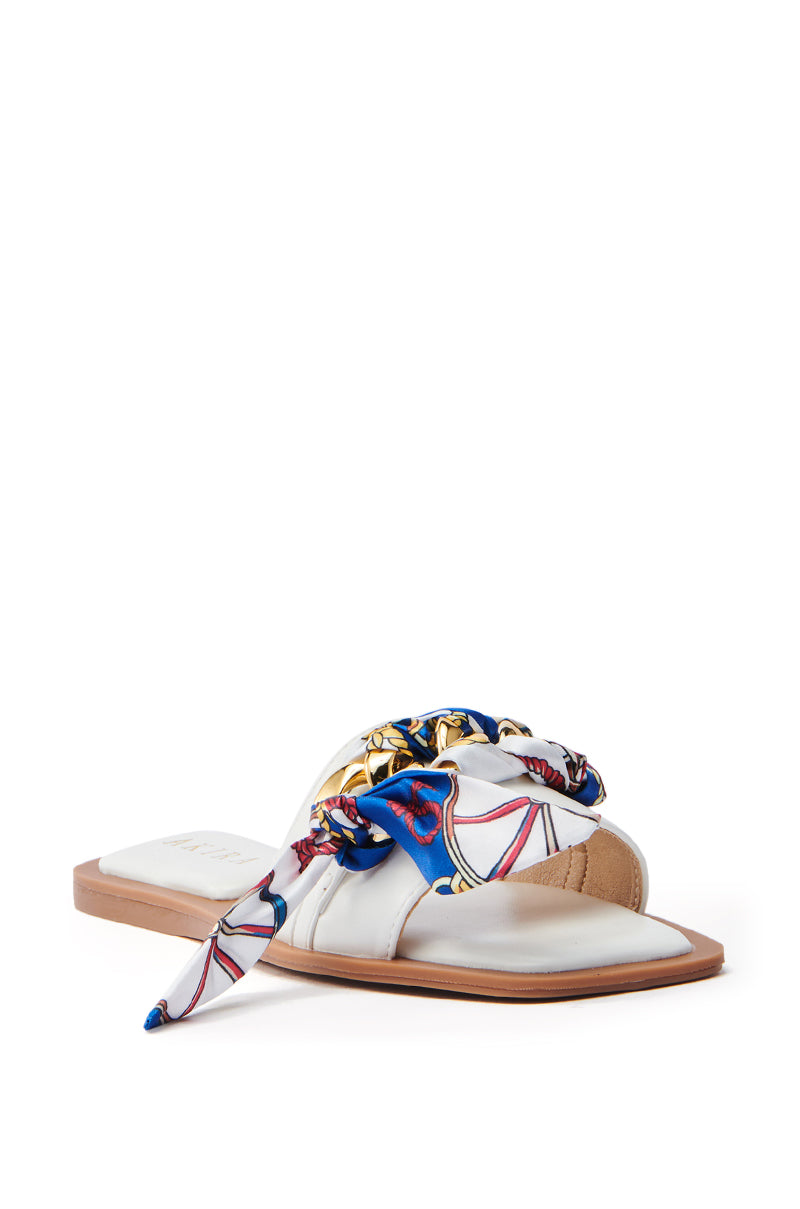 The AKIRA LORCAN WHITE SANDAL is a chic, stylish slide with a flat sole and easy slide-on fit. It features a white vegan leather base and a decorative fabric knot showcasing blue, red, and gold patterns.