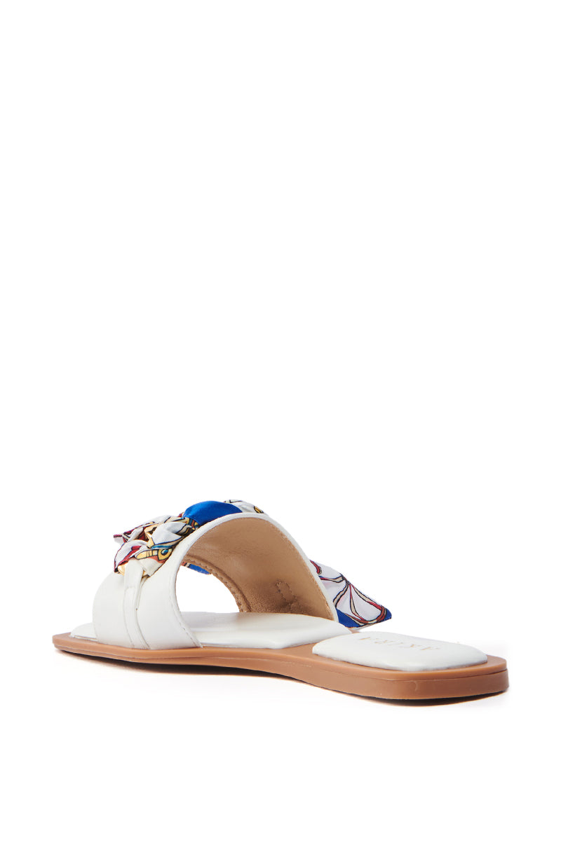 The AKIRA LORCAN WHITE SANDAL features a flat, tan sole with a white cushioned footbed. Its vegan leather woven strap showcases an intricate pattern with blue, red, and gold accents. Enjoy its easy slide-on fit for effortless style.