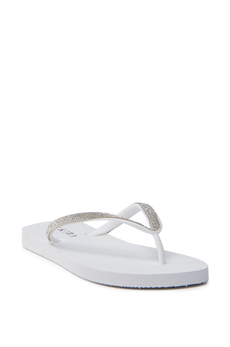 The AKIRA LORETA WHITE THONG FLIP FLOP features textured white straps with decorative rhinestones on a simple, flat sole. Made from durable PVC, it is displayed on a light background in a slightly angled side view.
